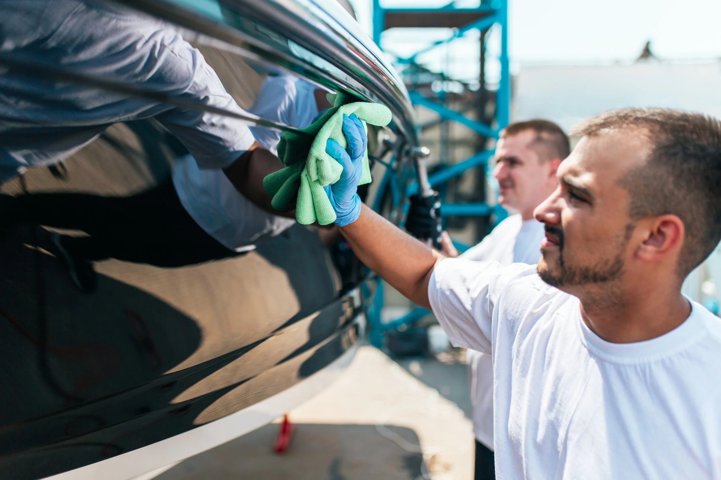 Two men inspecting a shiny black boat at a marina or boatyard. One man is cleaning or polishing the boat's surface with a cloth, while the other observes. Scaffolding and a clear sky are in the background.