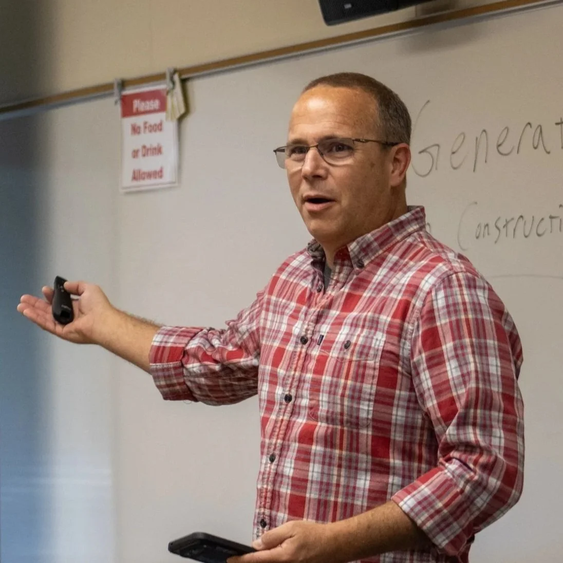 Man in glasses wearing a red plaid shirt giving a lecture in a classroom with a whiteboard behind him and a sign that says 'Please No Food or Drink Allowed'.