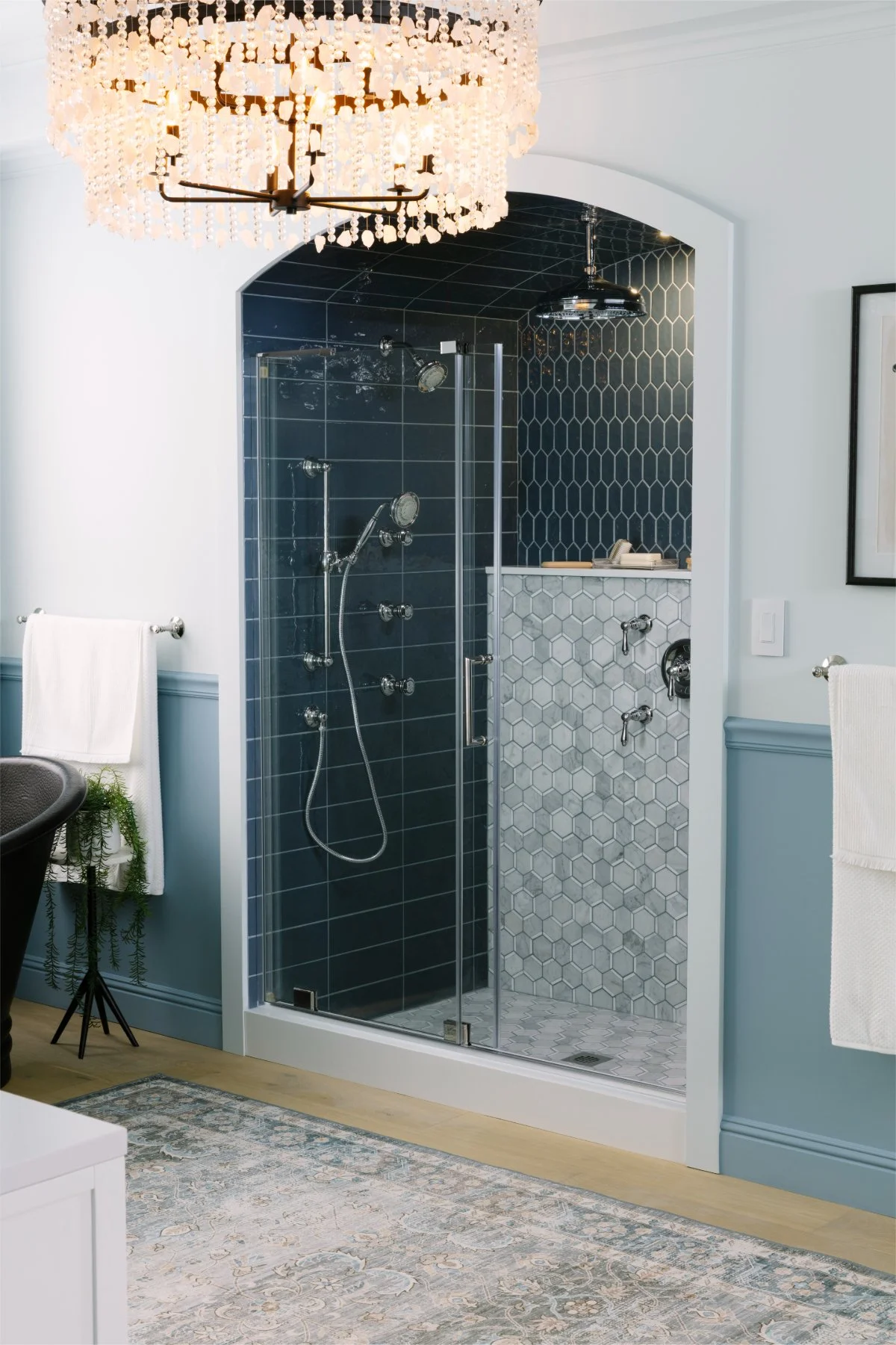 Modern bathroom featuring a walk-in shower with dark blue and white hexagonal tiles, decorated with a chandelier hanging from the ceiling, and light blue walls with a white baseboard.