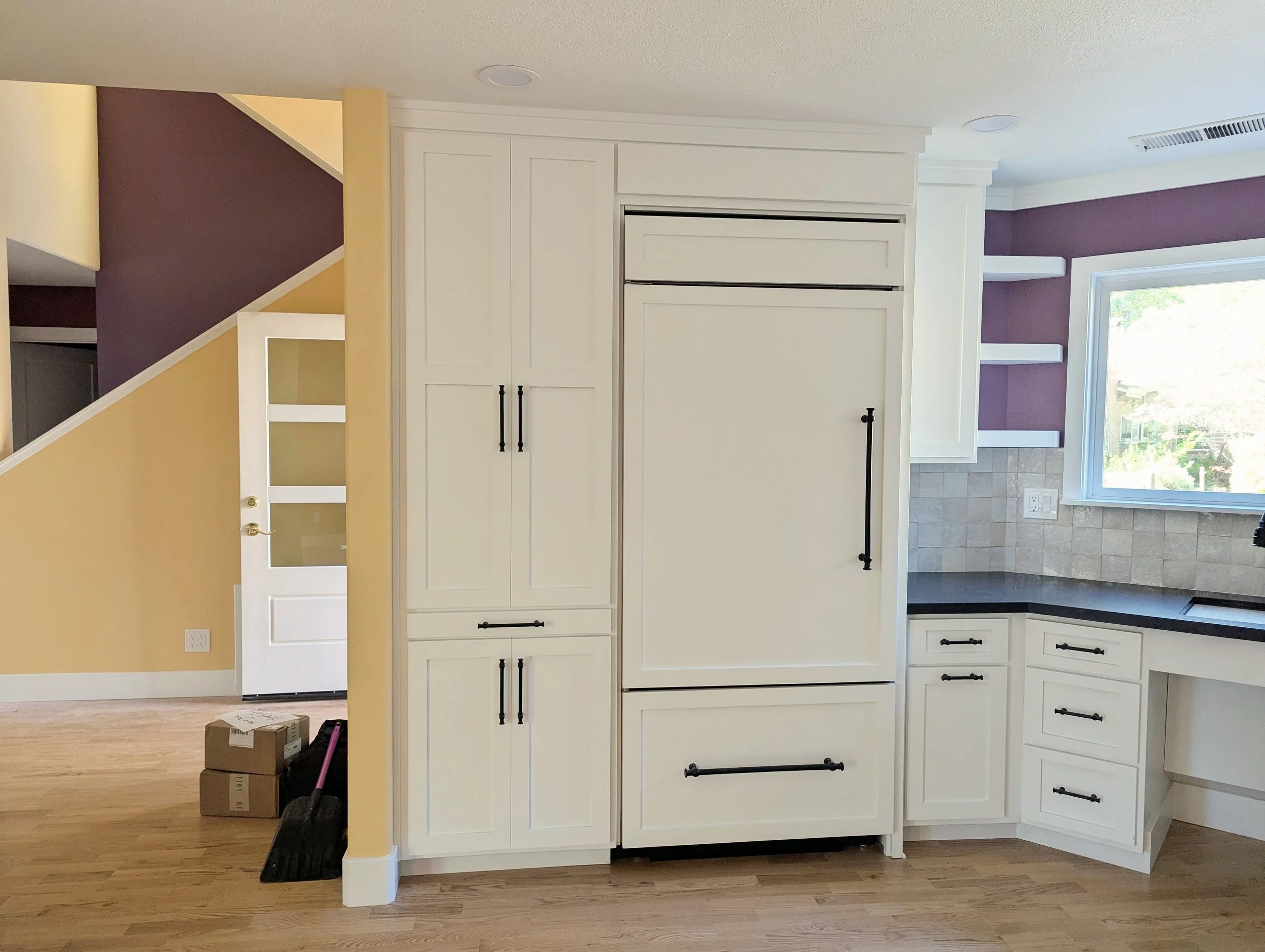 Bright kitchen with white cabinets, large window letting in natural light, and purple accent wall. Some moving boxes and cleaning tools on the wood floor.