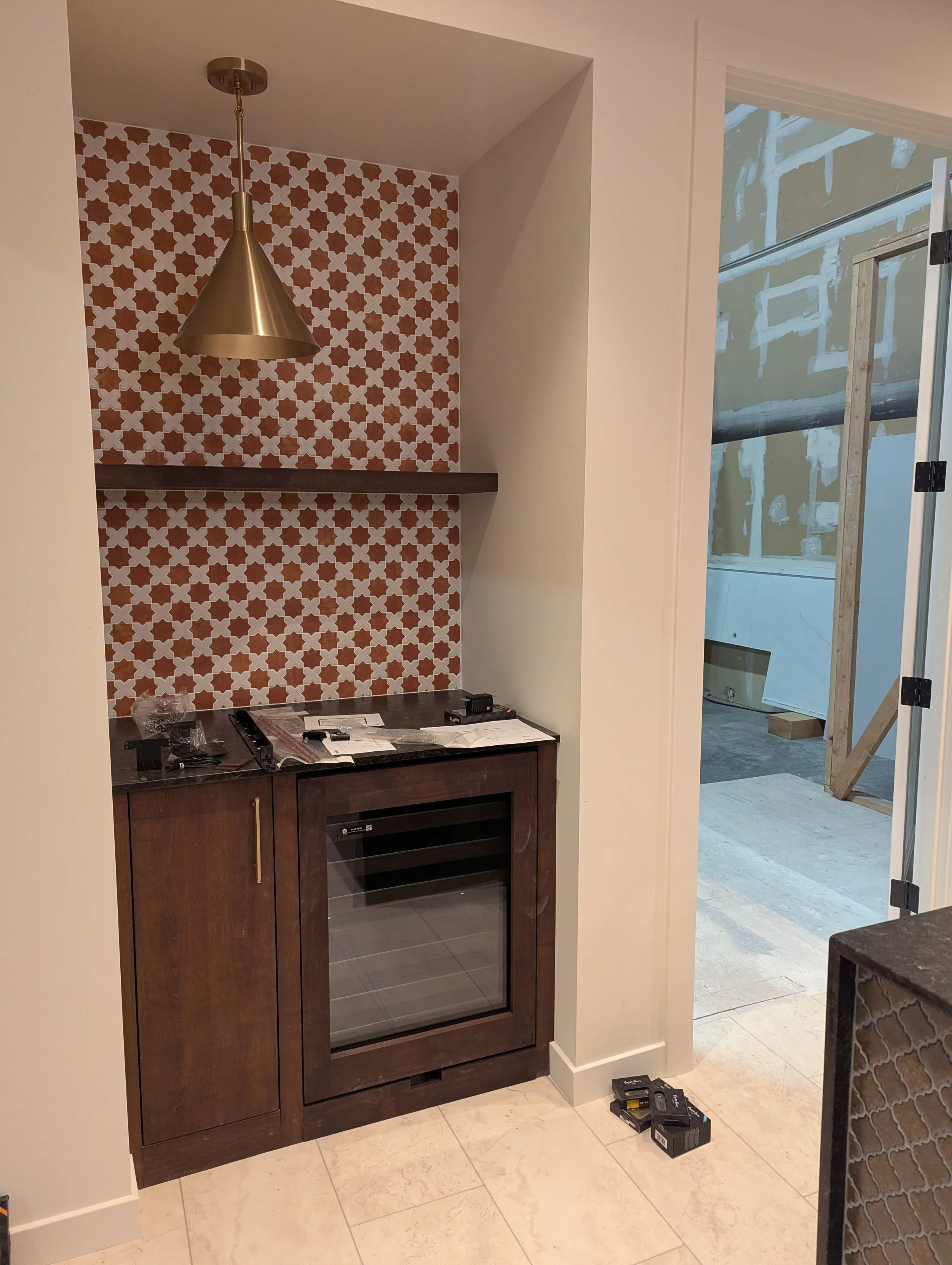 Kitchen or bar area under construction with a dark wood cabinet, a mini fridge, a decorative red and white tile backsplash, and a brass pendant light. There are construction materials and tools on the floor and countertop, and a doorway leading to an