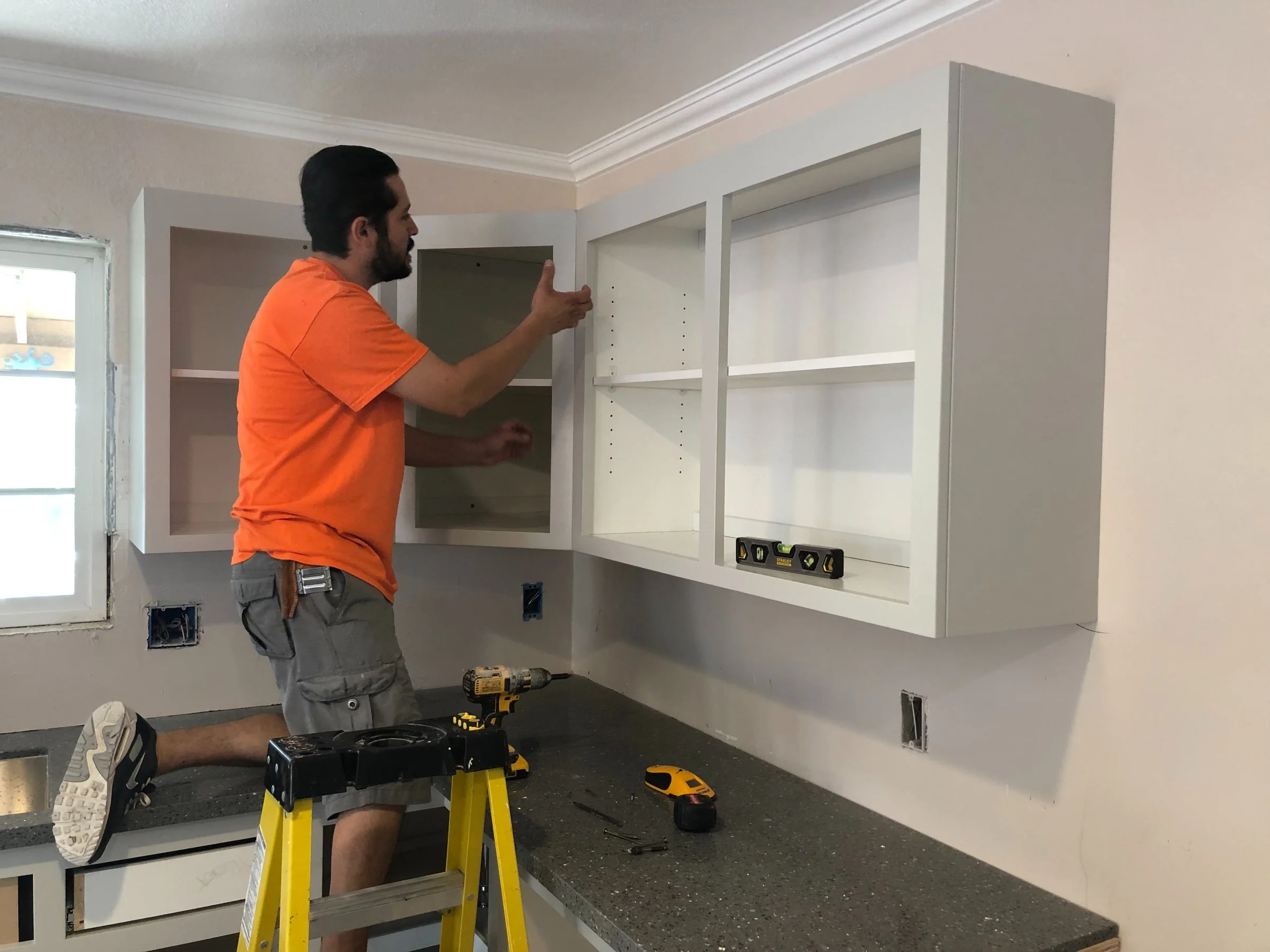 A man wearing an orange t-shirt and gray shorts is standing on a yellow step stool while installing white upper kitchen cabinets. He is using a drill on the cabinet. The room has white walls and a window on the left side.