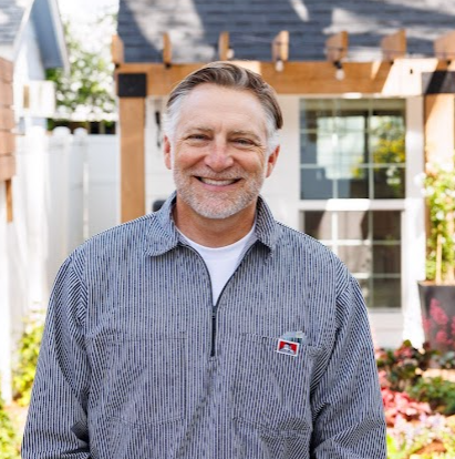 Smiling middle-aged man standing outdoors in front of a house with a garden and a wooden pergola.