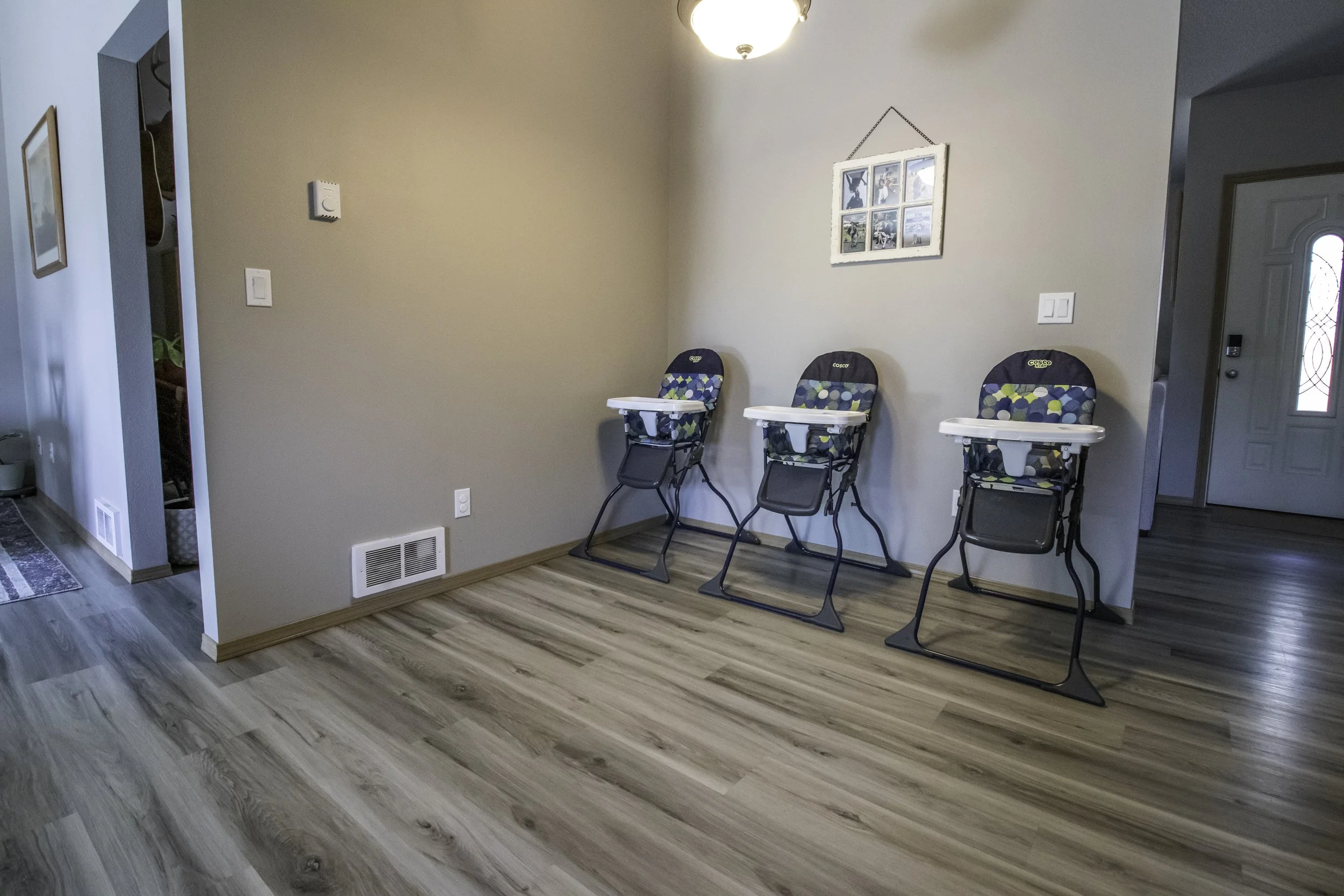 Three baby high chairs with colorful patterned fabric seats are lined up against a beige wall in a home interior, with a framed collage of photos hanging above them.