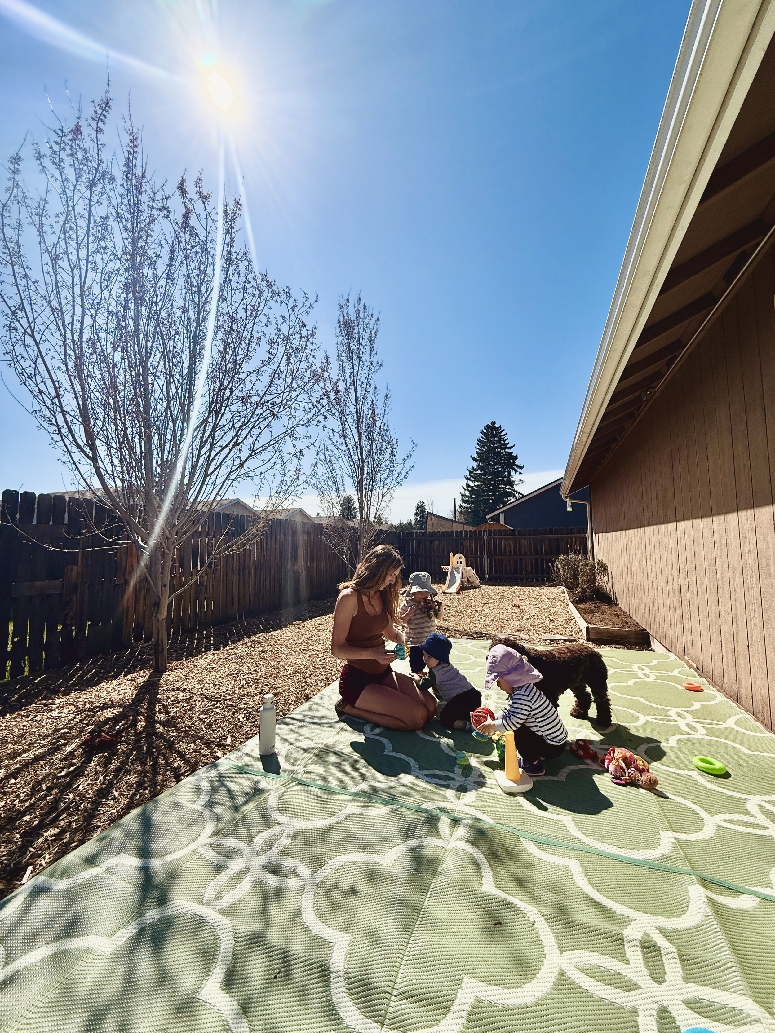 A woman with three children and a black dog in a backyard, playing on a mat under a bright sunny sky with a wooden fence and trees in the background.