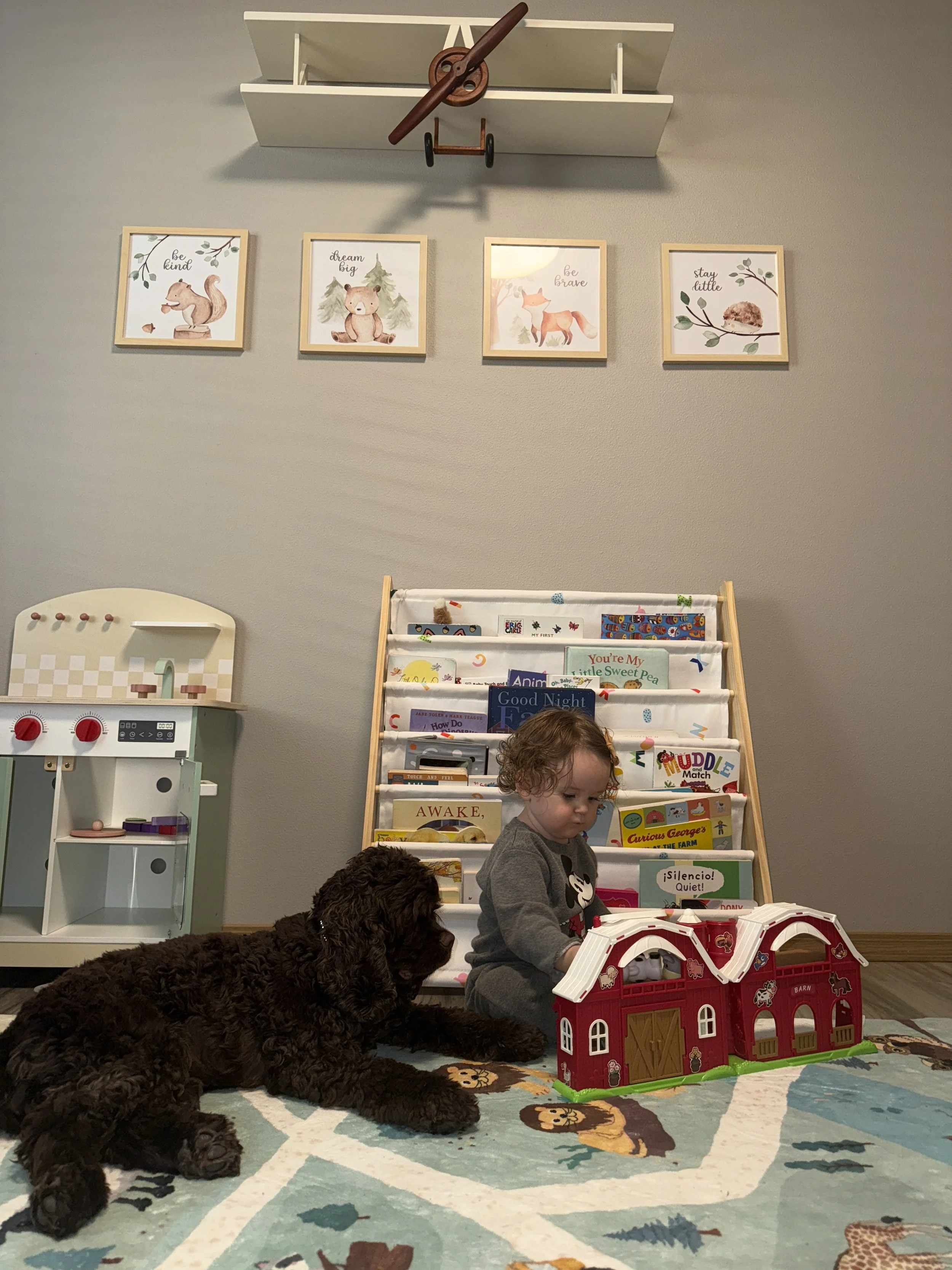 A young girl and a black puppy playing on a children's rug in a playroom, with a red toy barn and a book rack in the background, decorated with framed animal-themed artwork on the wall.