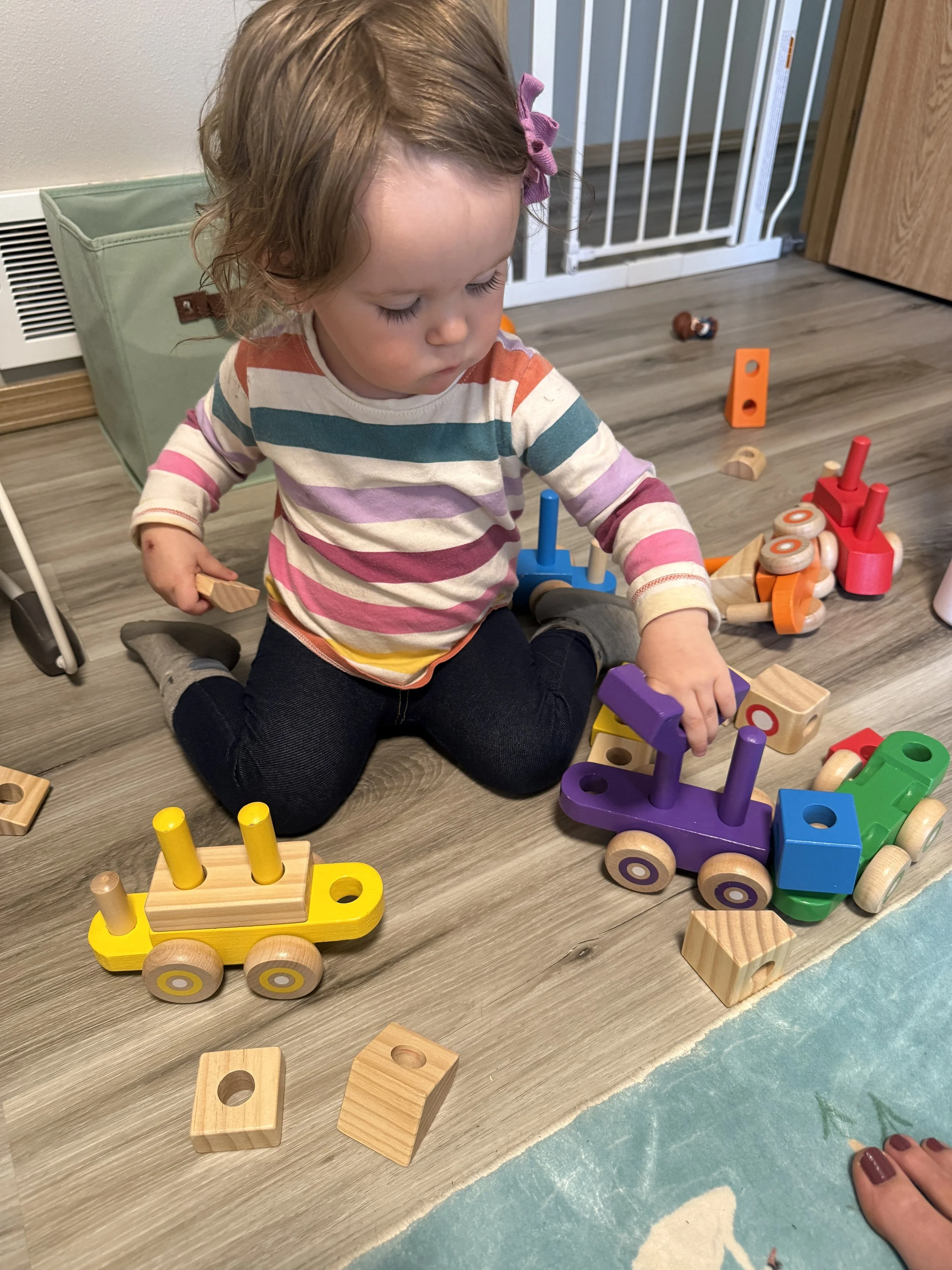 A young girl playing with colorful wooden toy trains and blocks on a wooden floor.