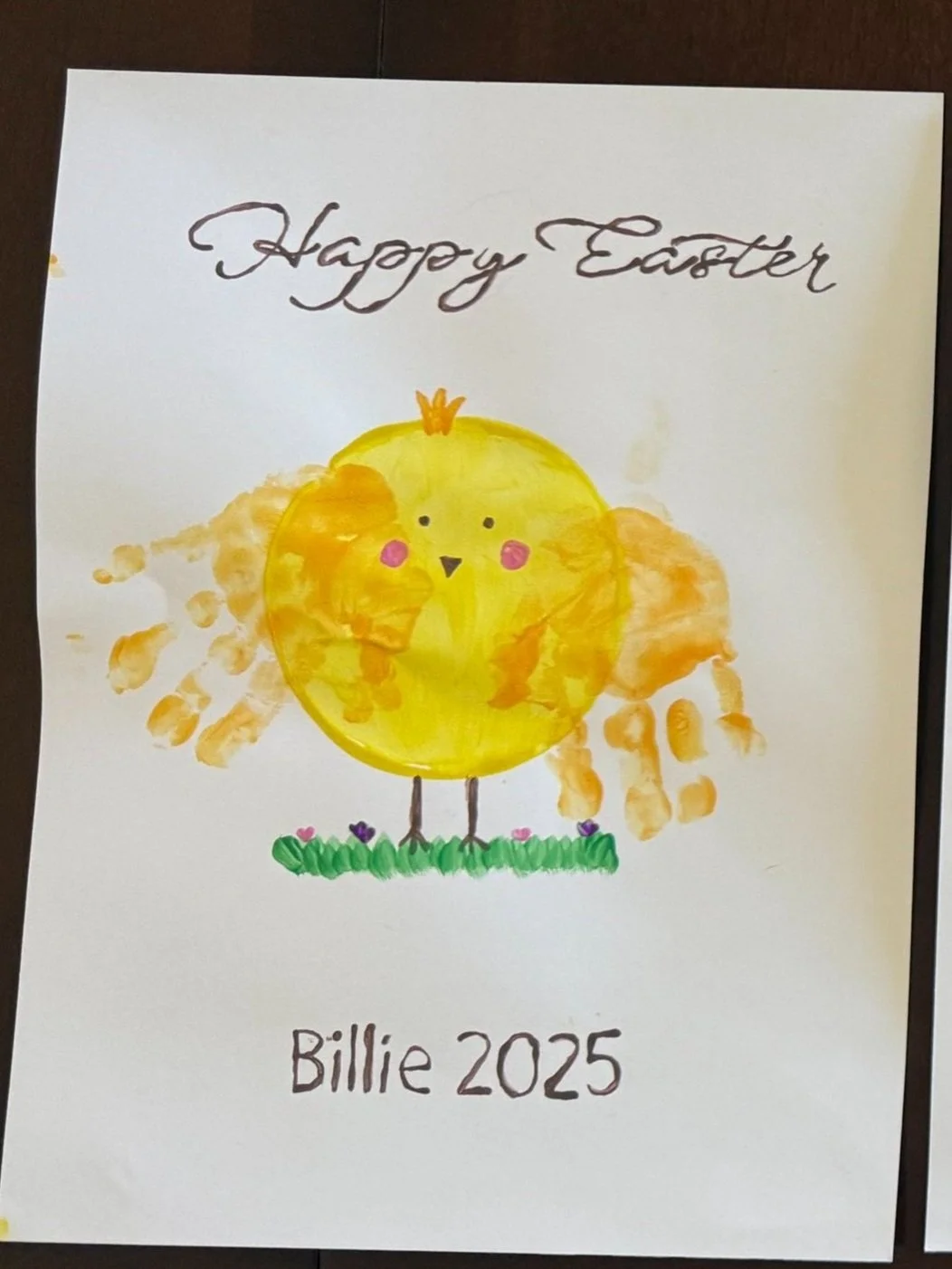 Handmade Easter card featuring a chick with a yellow body, pink cheeks, and little legs, standing on green grass with small flowers, with "Happy Easter" at the top and "Billie 2025" at the bottom.