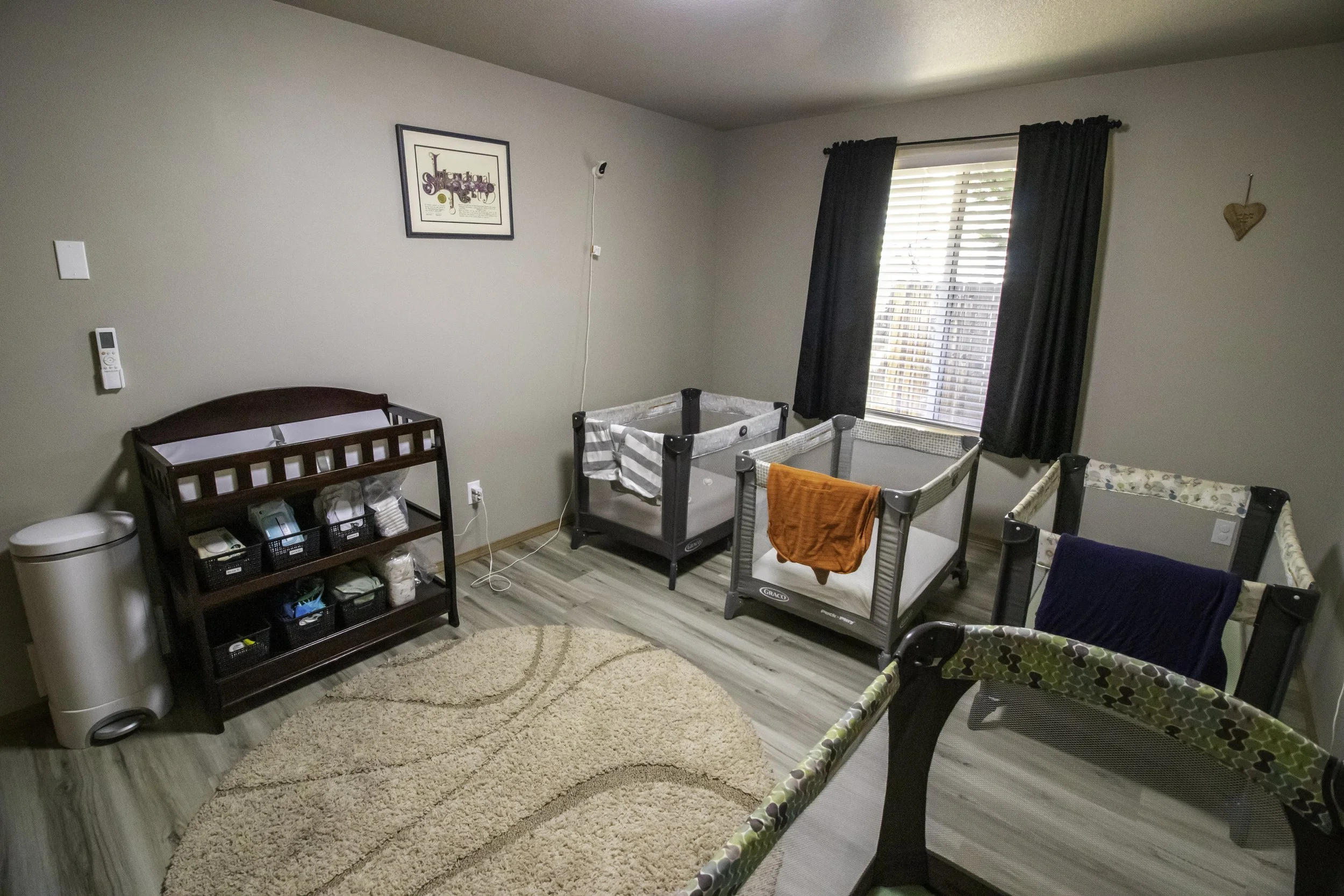 Bedroom with light-colored walls, wood floor, four cribs with blankets, a beige rug, a window with blinds and curtains, a framed picture on the wall, a small dark wood changing table with baskets and items, and a white odor control unit beside it.
