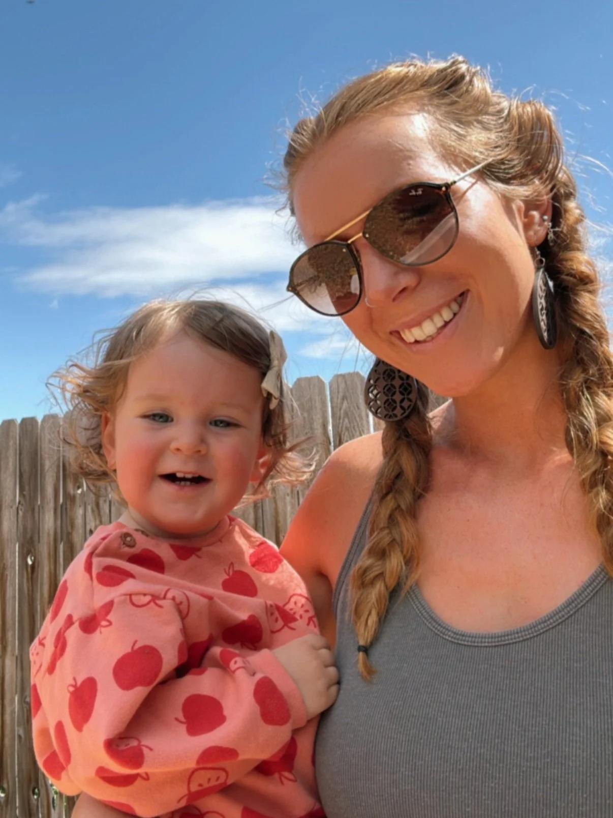 A woman with braided hair and sunglasses smiling while holding a young girl in front of a wooden fence and blue sky.