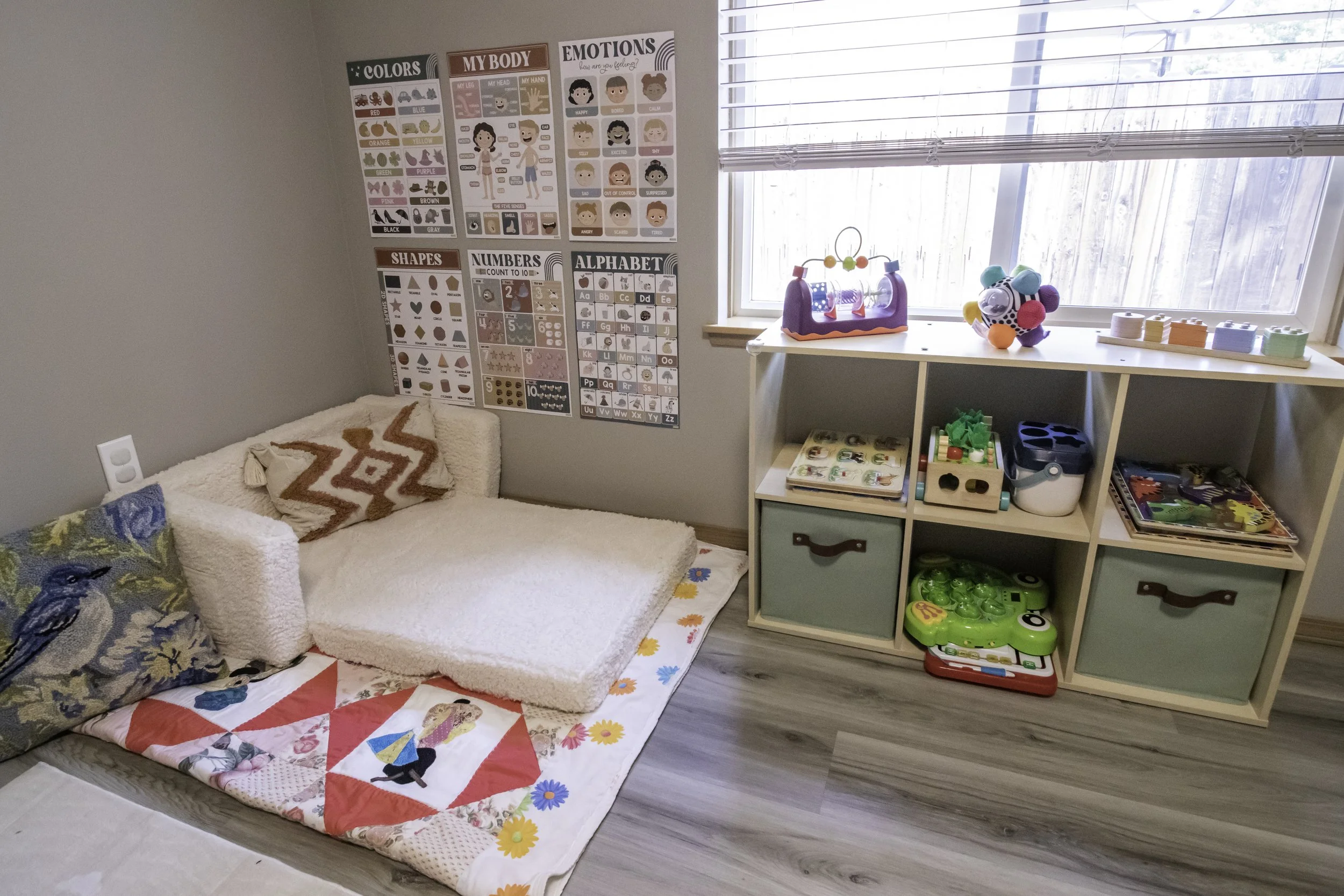 A cozy children's playroom with educational posters on the wall, a soft bed with colorful pillows and a quilt, a window with blinds, and a storage unit filled with toys and books.
