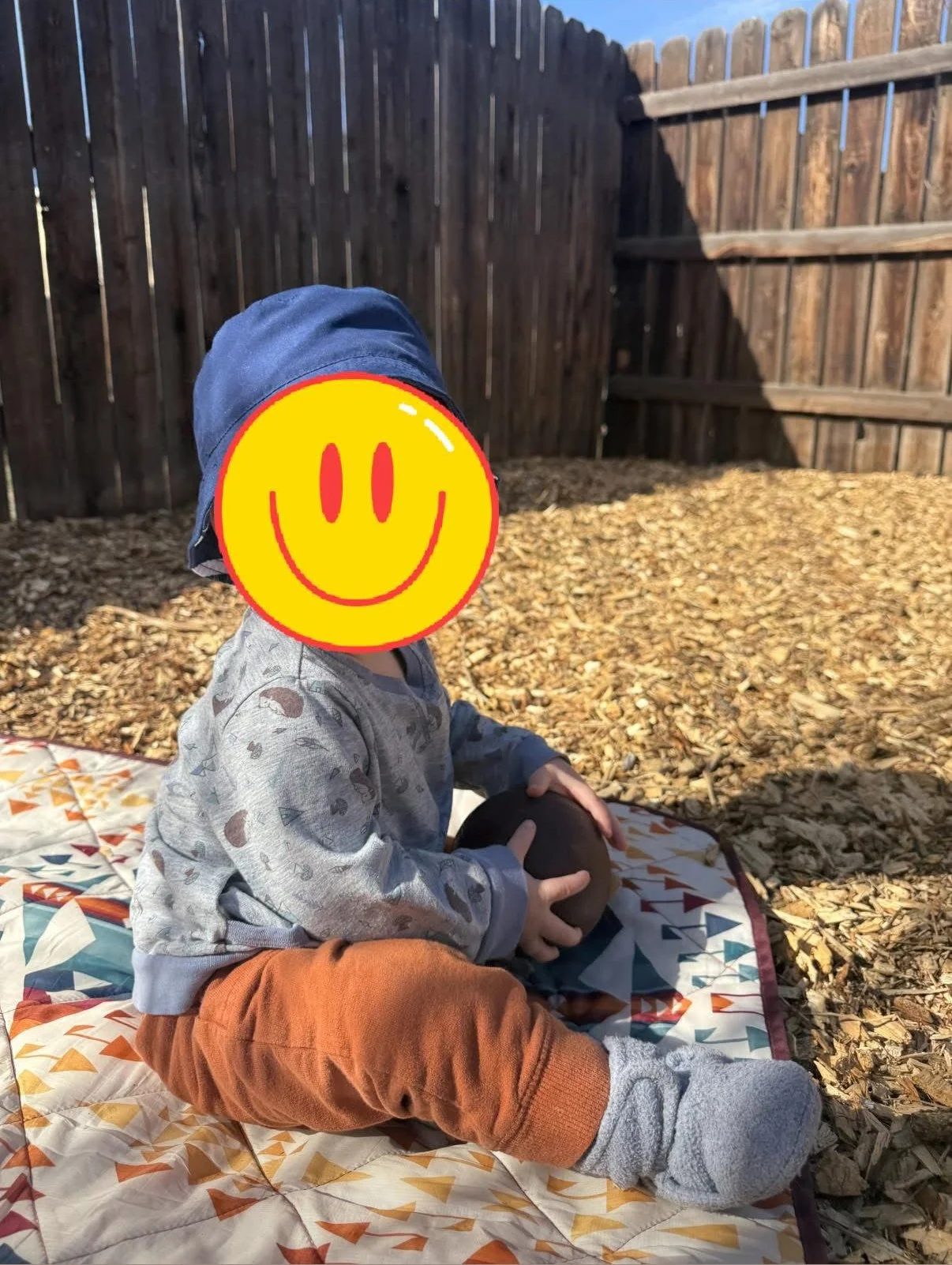 Child sitting outdoors on a quilted blanket, holding a round object, with a wooden fence in the background, in sunlight.