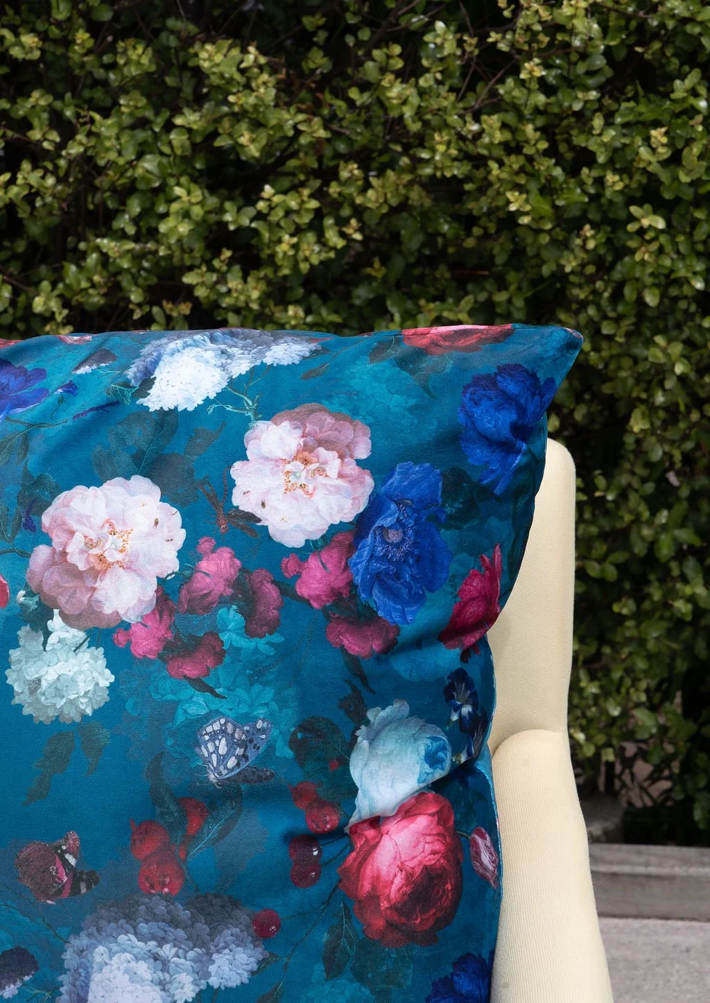 Close-up of a blue floral pillow with pink, white, blue, and red flowers and butterflies, resting on a cream-colored chair, with a green bush in the background.