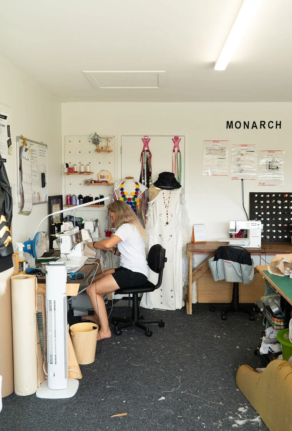 Chelsea working on a sewing machine in a workspace with dress forms, fabric rolls, and sewing supplies.