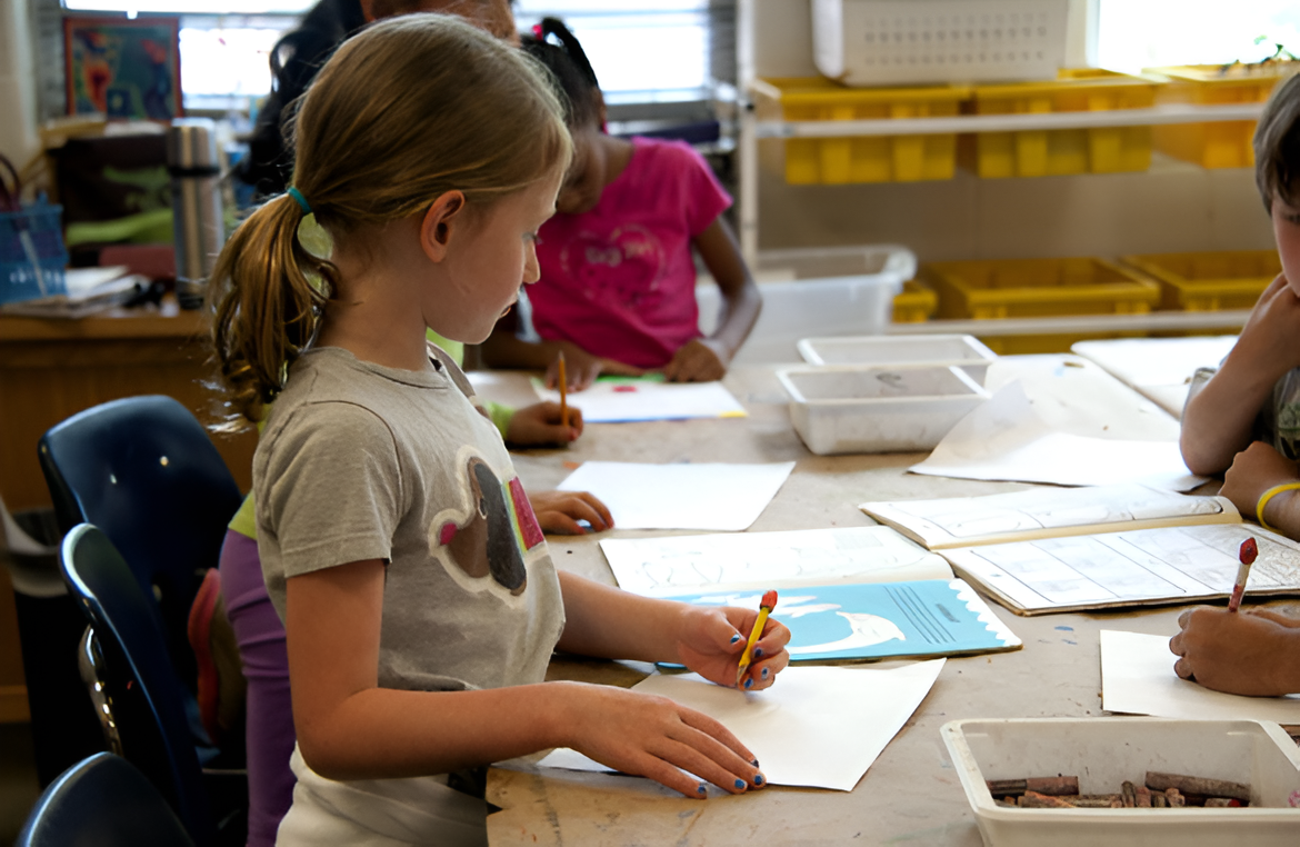 Children sitting at a table engaged in drawing or writing activities in a classroom.