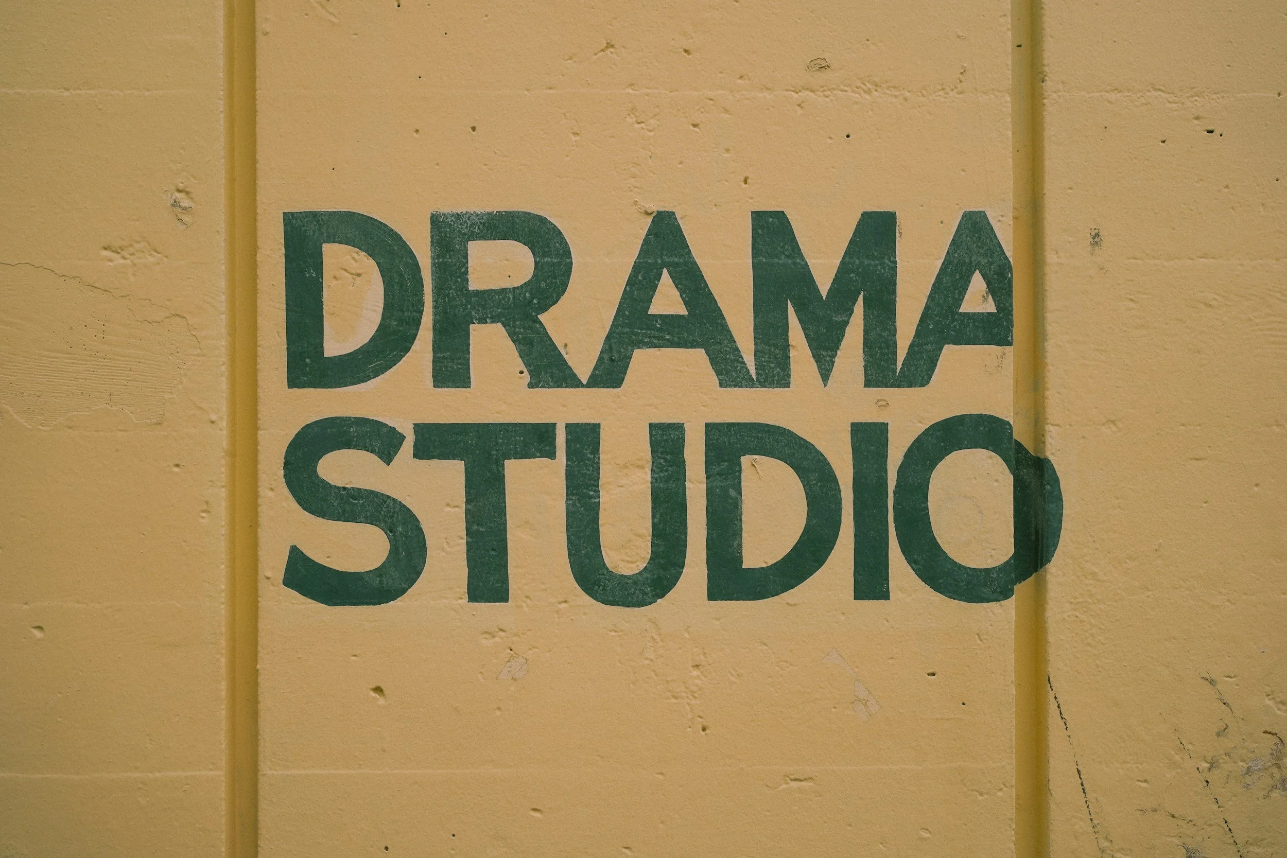 Close-up of a yellow wall with peeling paint and a green sign that reads 'DRAFTING STUDIO'.