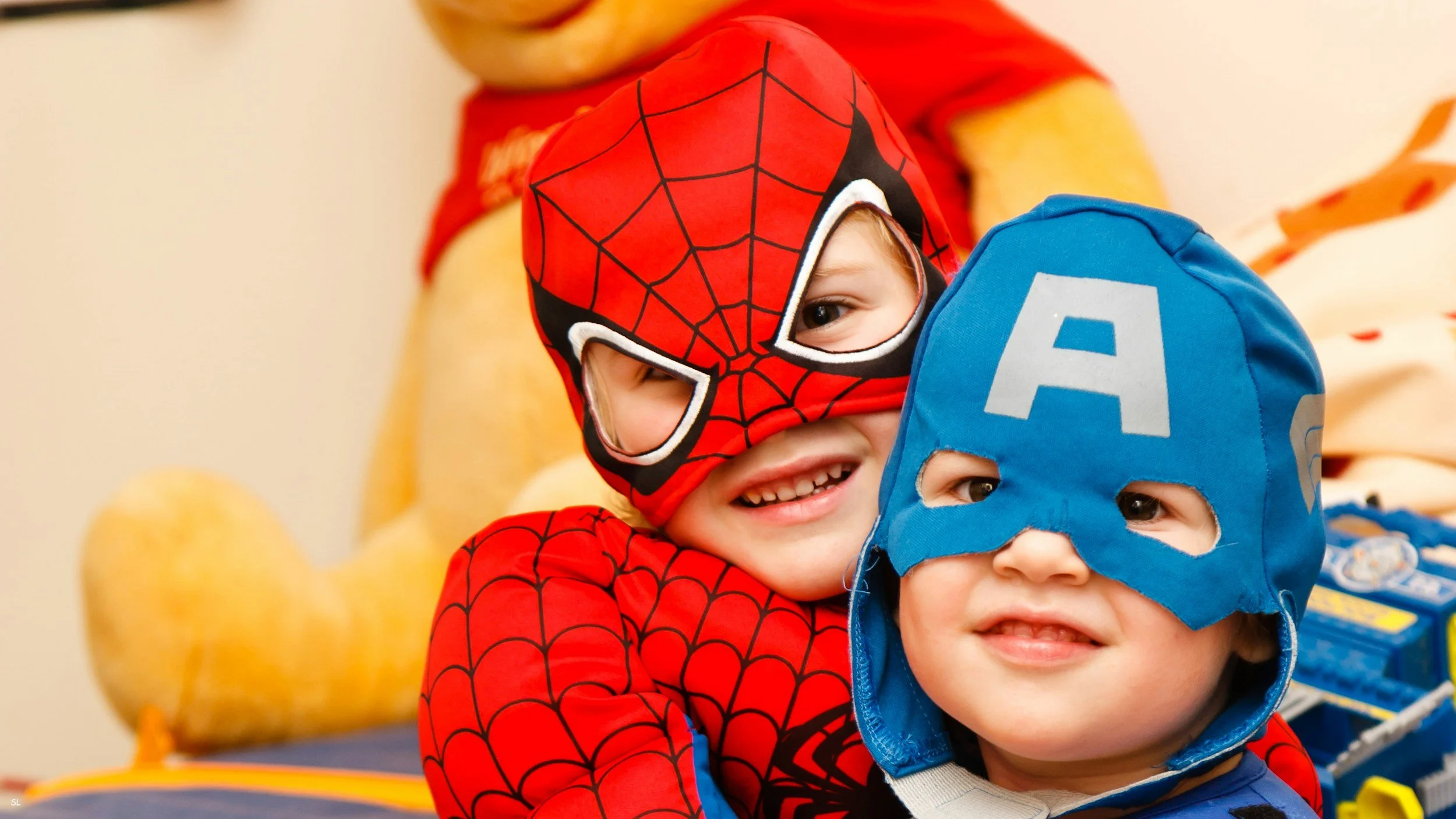 Two children in superhero costumes jumping and playing indoors, with a child dressed as Spider-Man and another as Captain America.