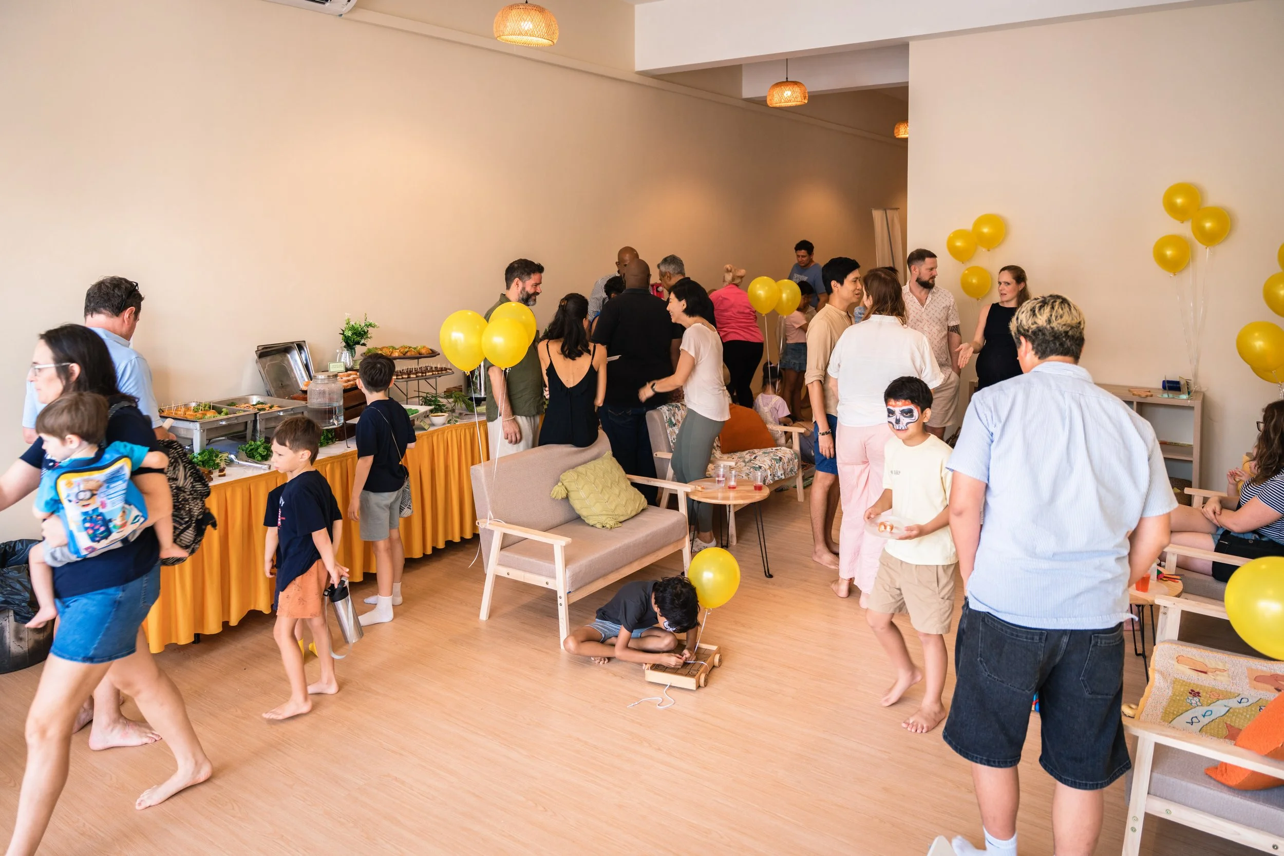 A lively indoor party with children and adults mingling, balloons, and a table with food. People are standing, talking, and playing; some children are on the floor. The room has warm lighting from hanging lamps and a beige wall background.