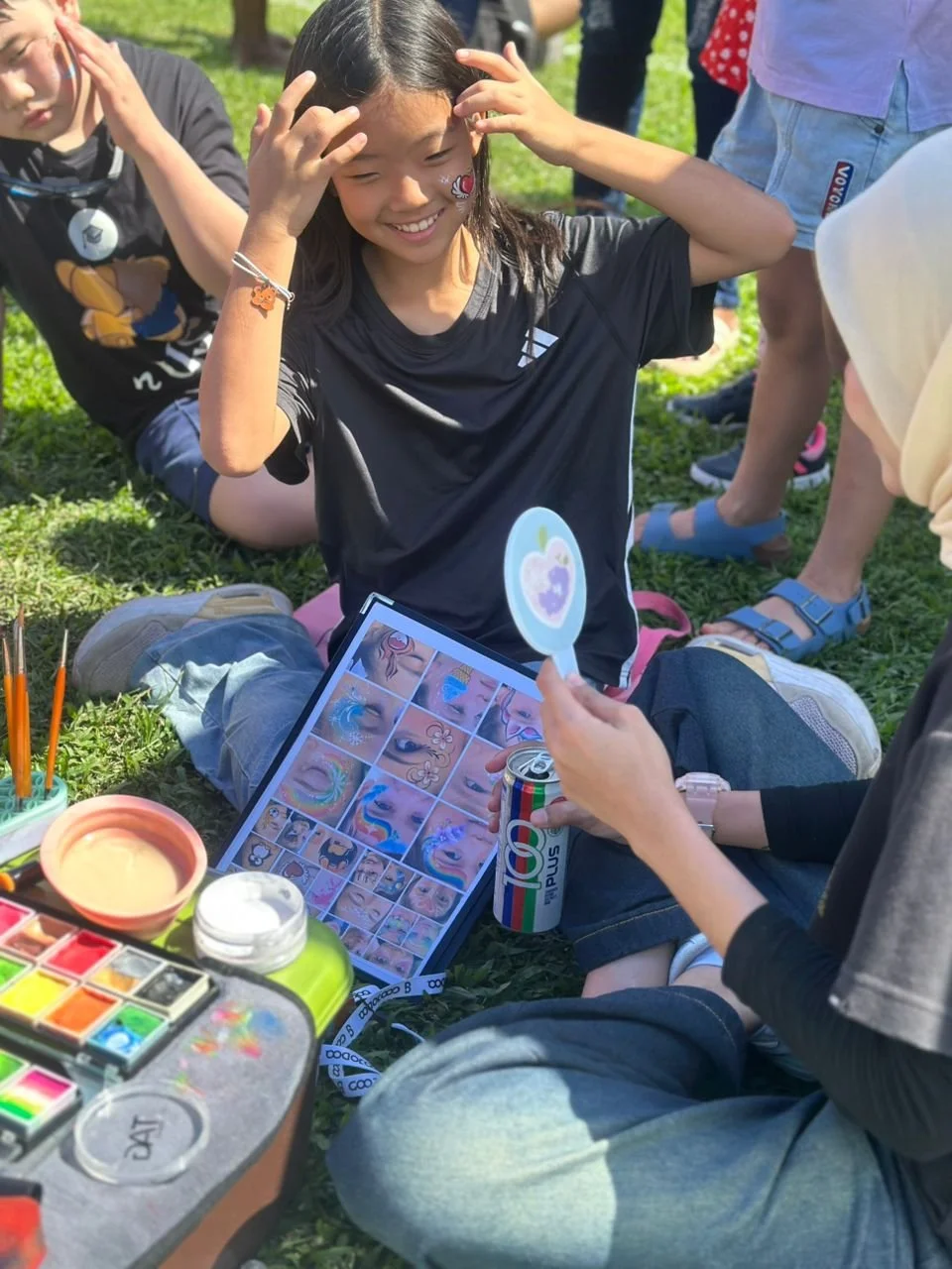 A girl sitting on grass gets a face painted at an outdoor event, smiling with her hands on her head, while an artist paints her face. Art supplies and reference images are visible nearby.