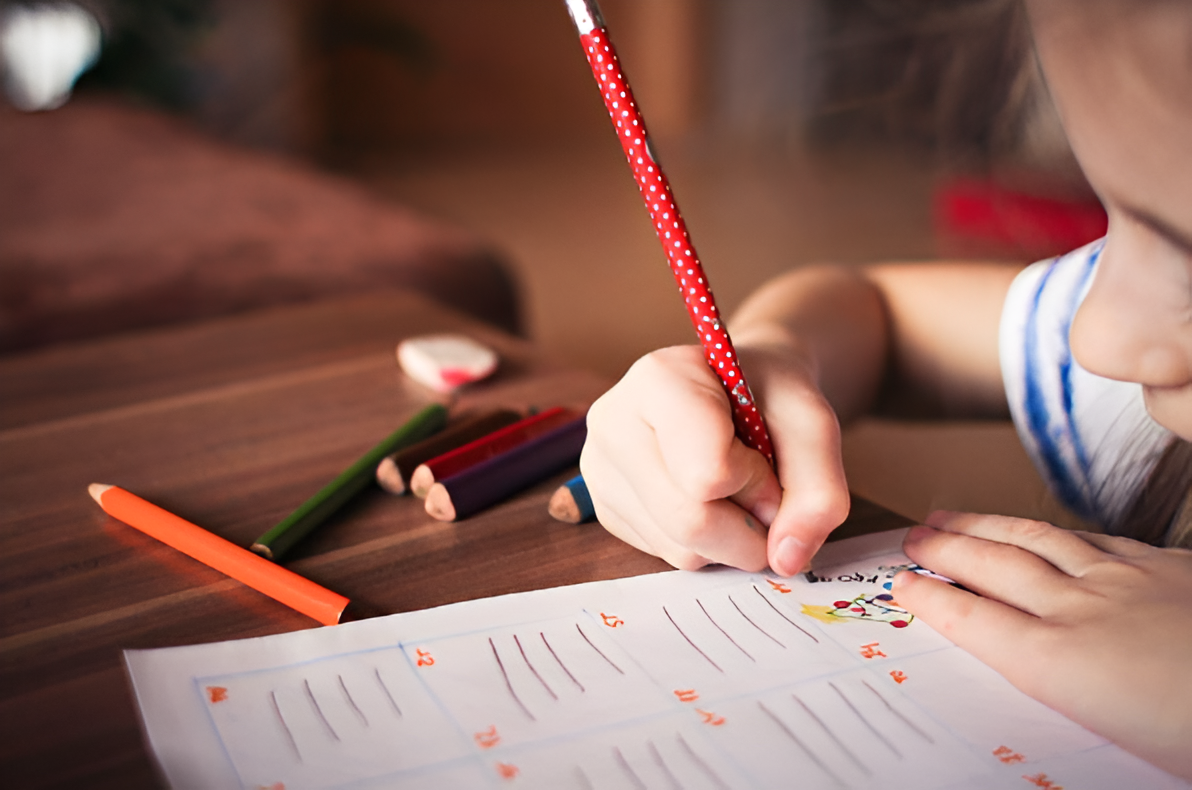 A child writes on a sheet of paper with colorful pens on a wooden table.