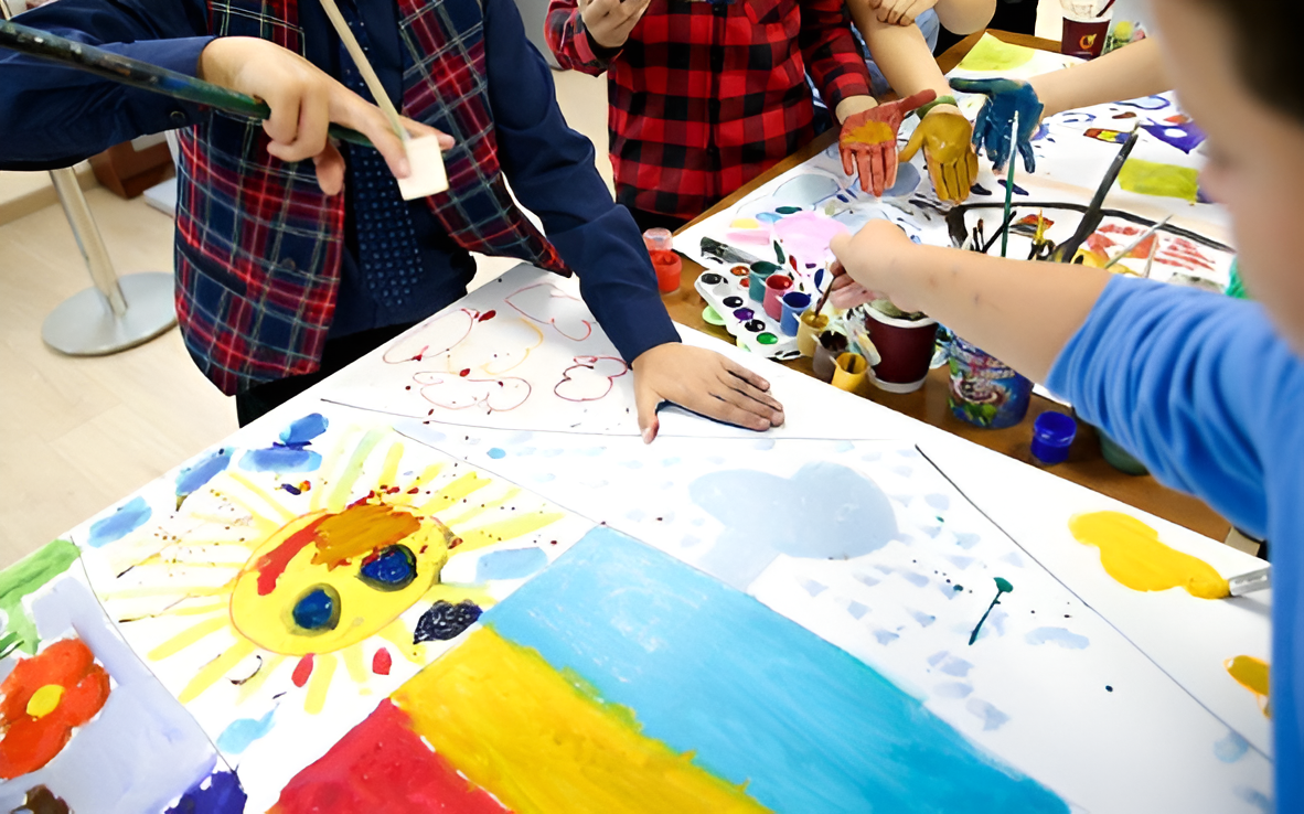 Children are painting and drawing on large sheets of paper with colorful paints and brushes in an art class.