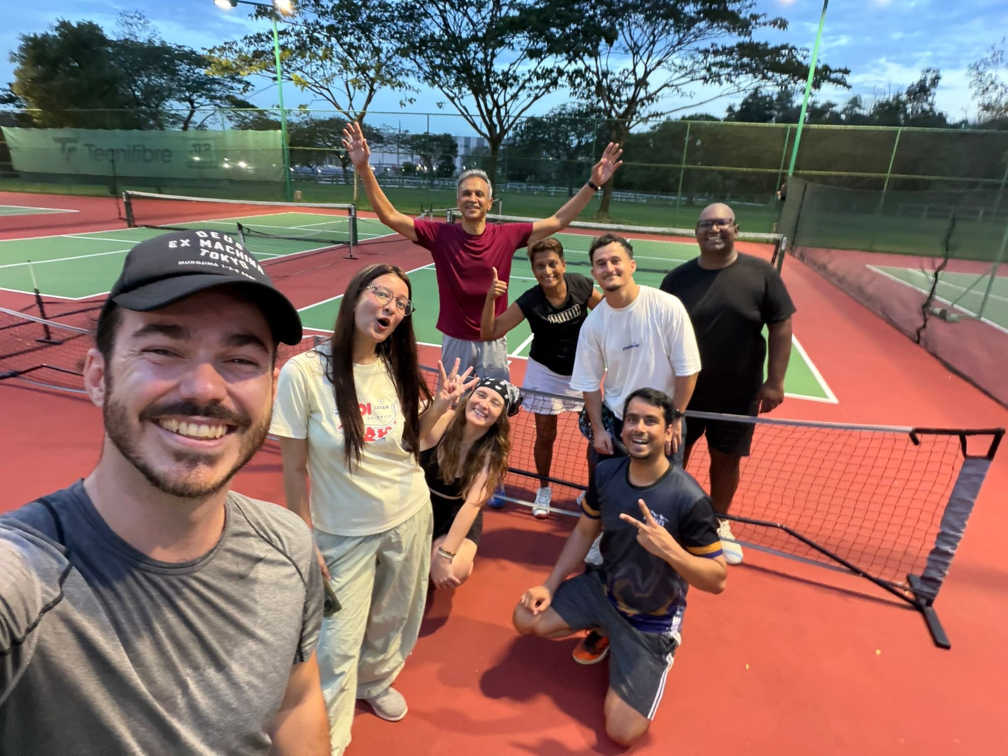 Group of eight people taking a selfie on a tennis court during the evening. They are smiling and making various hand gestures. The court is surrounded by trees and fencing, with a sky that is transitioning from day to night.