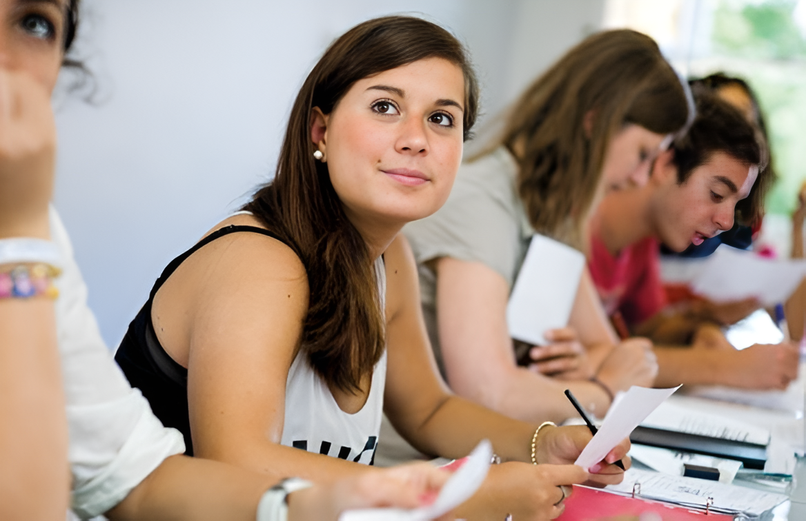Young woman with long brown hair smiling while sitting at a table with others, holding a smartphone in her hand, in a classroom or meeting setting.