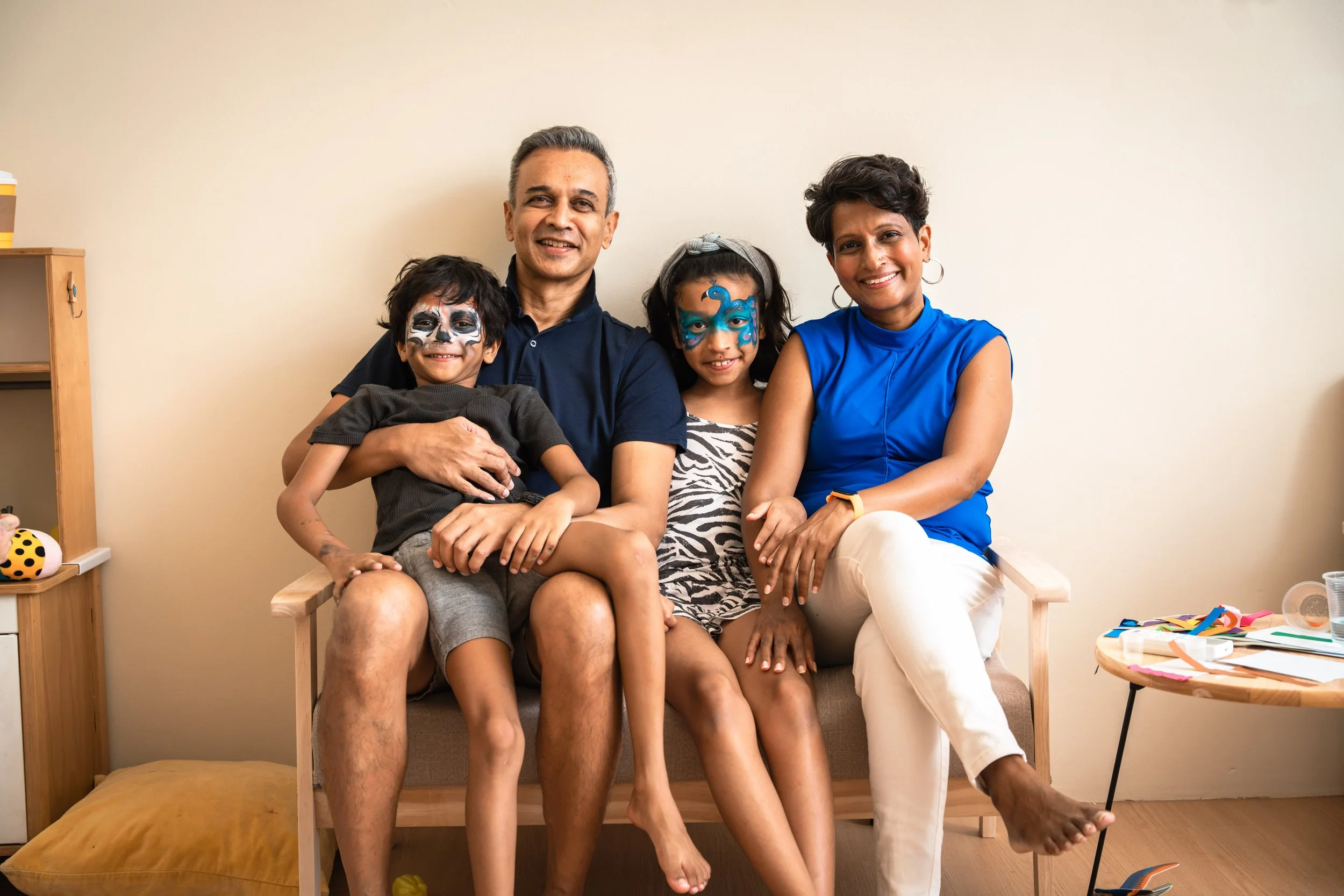 A family of four sitting together on a bench, with face paint on their faces. The father is smiling with his arm around his son, who has skeleton face paint. The daughter has butterfly face paint, and the mother is smiling, wearing hoop earrings and a blue top.