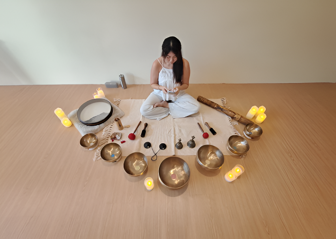A woman sitting cross-legged on a beige mat, surrounded by singing bowls, candles, and musical instruments, with a plain wall background.