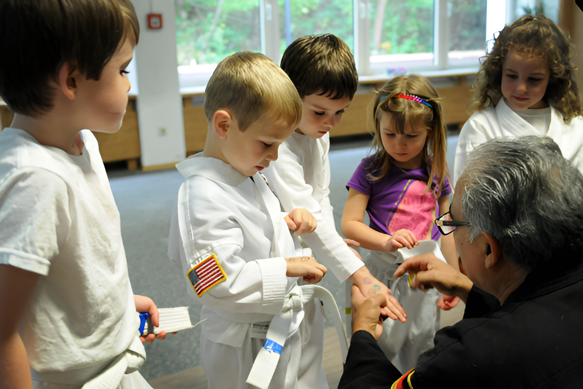 Children in martial arts uniforms receiving assistance from an instructor in a classroom.