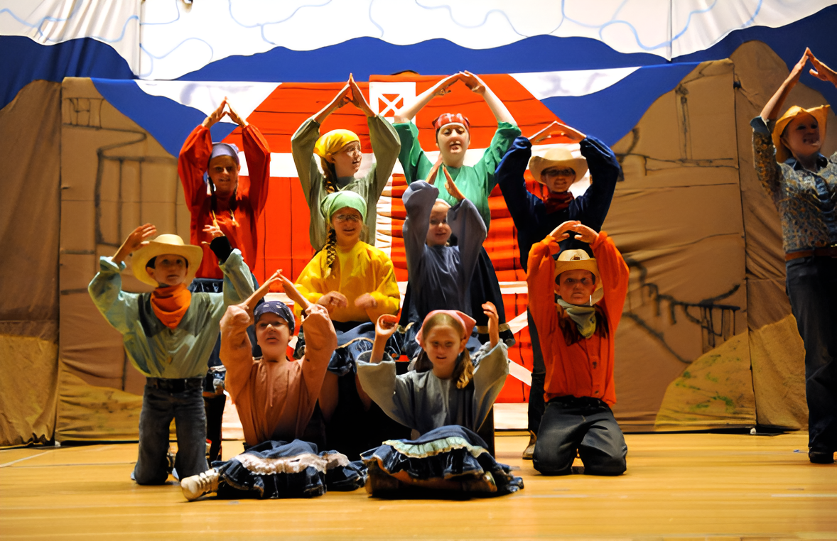 Children in cowboy costumes performing on stage with a backdrop of a barn and mountains.