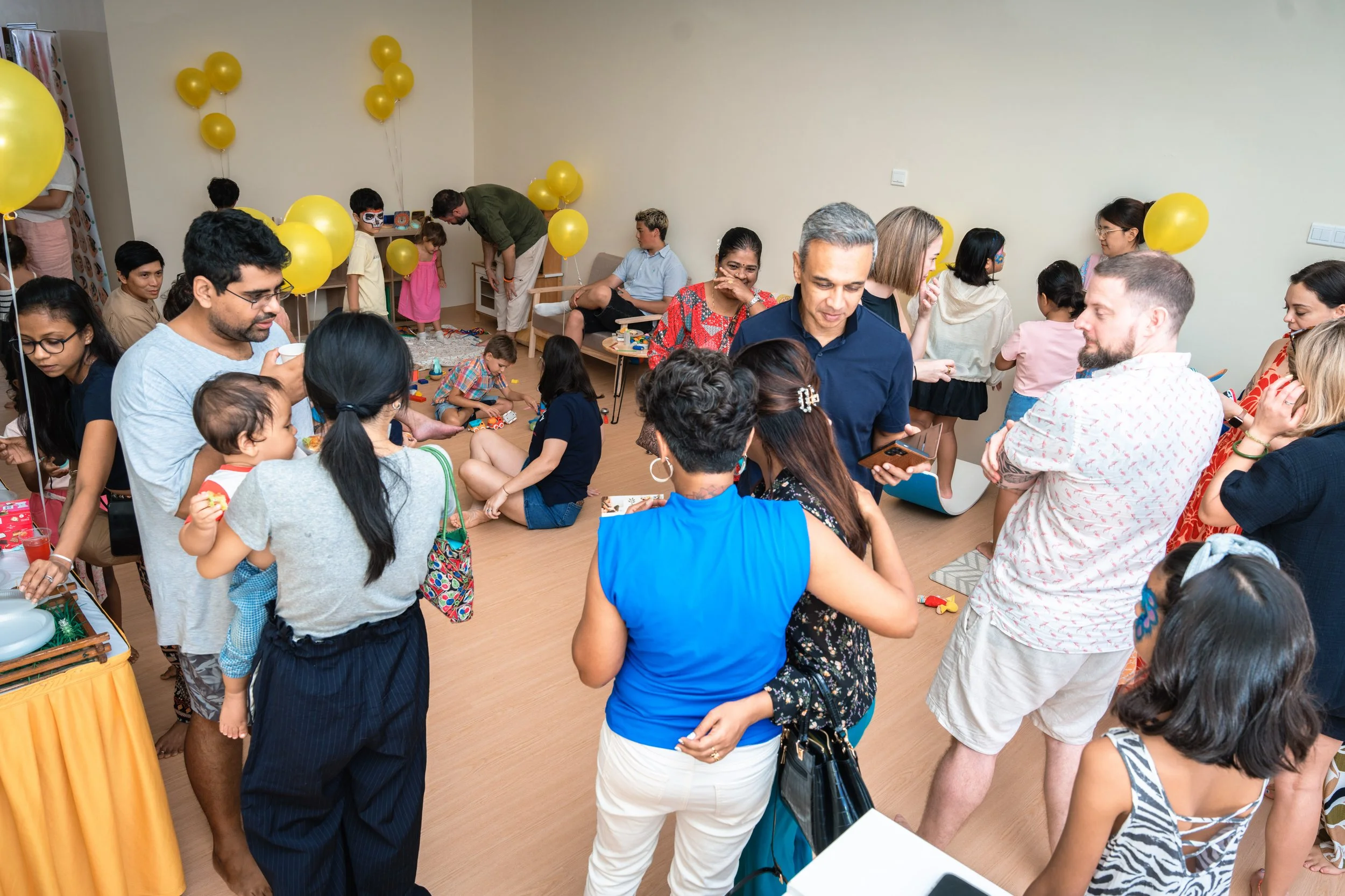 A crowded indoor birthday party with children and adults, yellow balloons, and a table with party items.