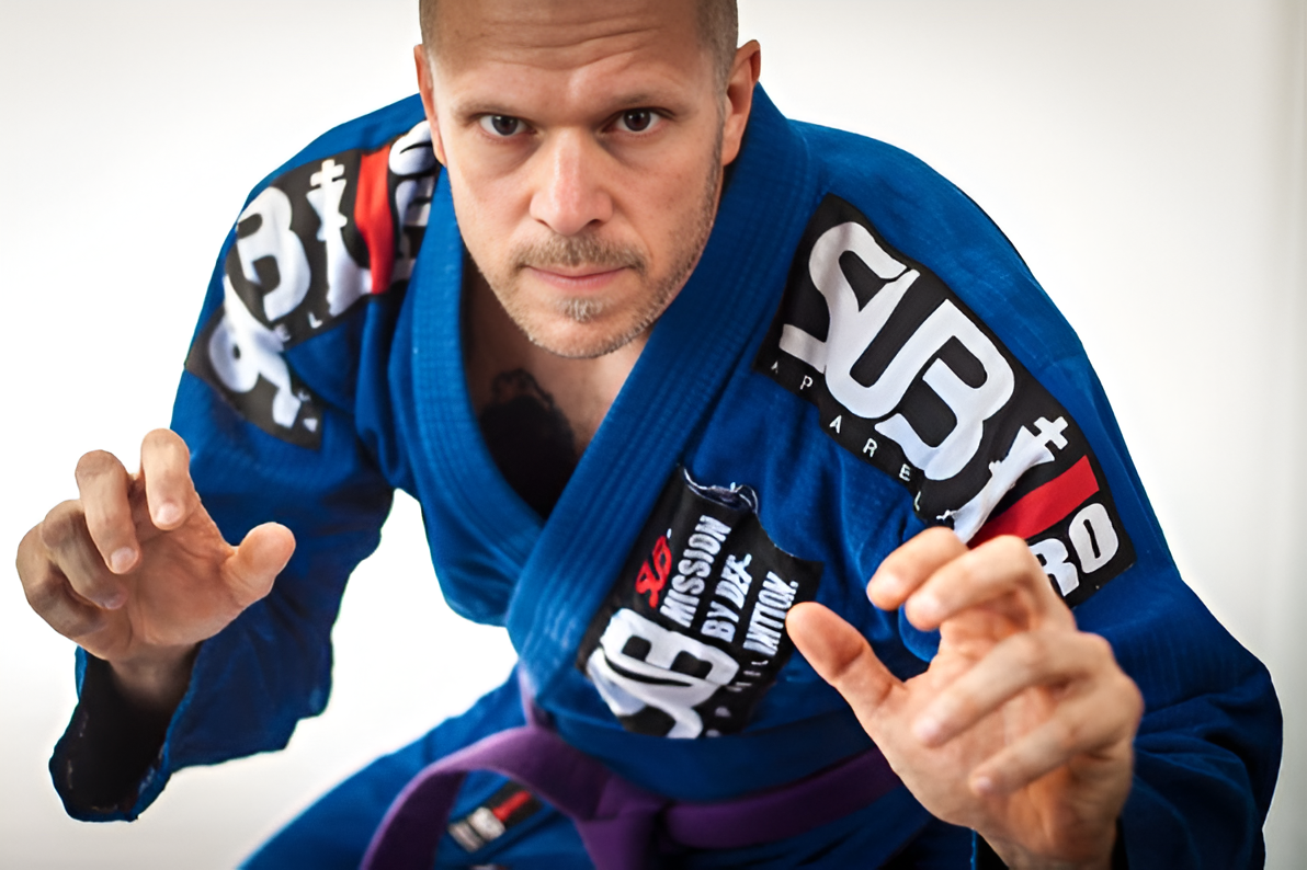 A man in a blue Brazilian Jiu-Jitsu gi is in a fighting stance, looking directly at the camera, with a focused expression.