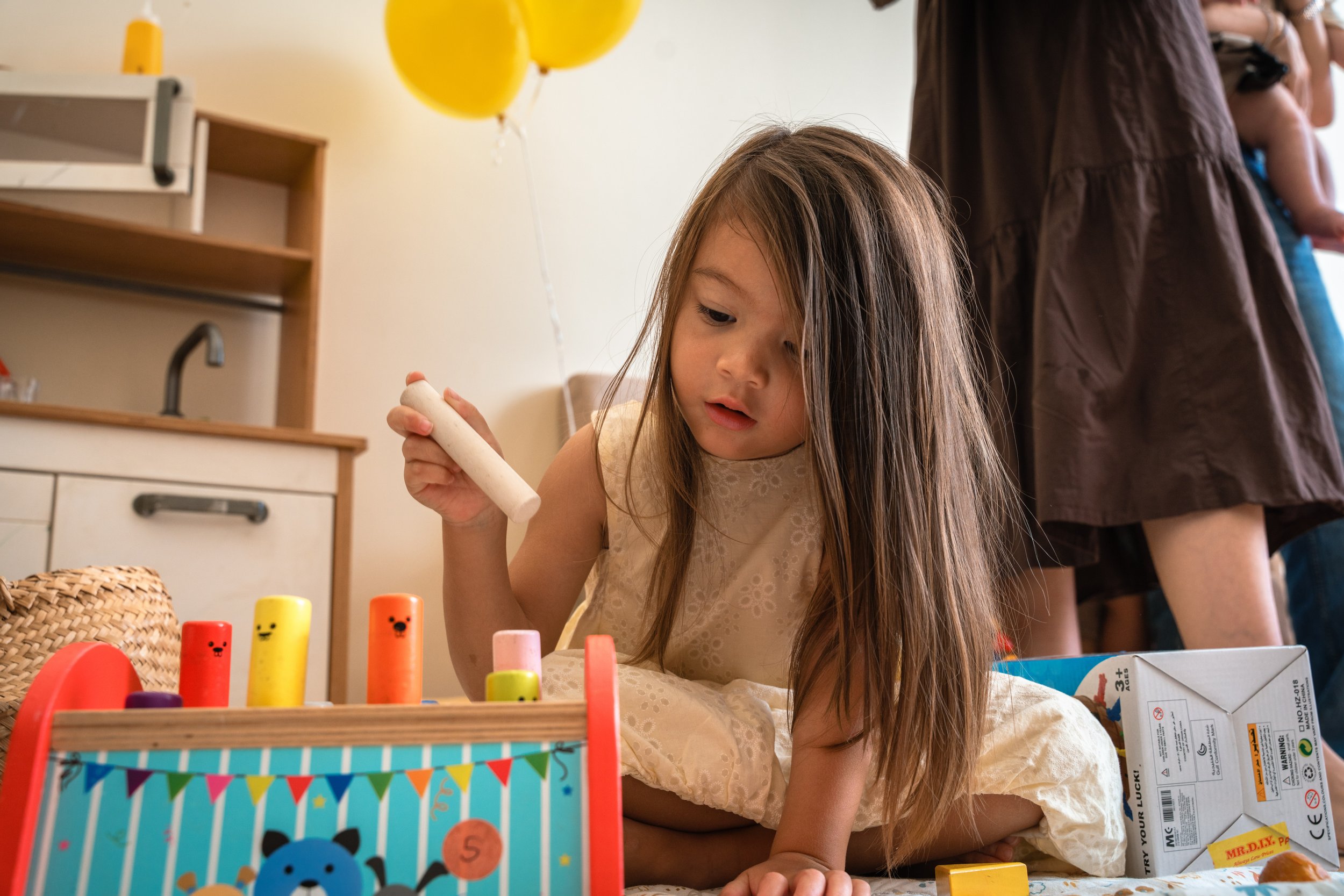 A young girl with long brown hair playing with colorful stacking toys at a birthday party. A basket and balloons are visible in the background.