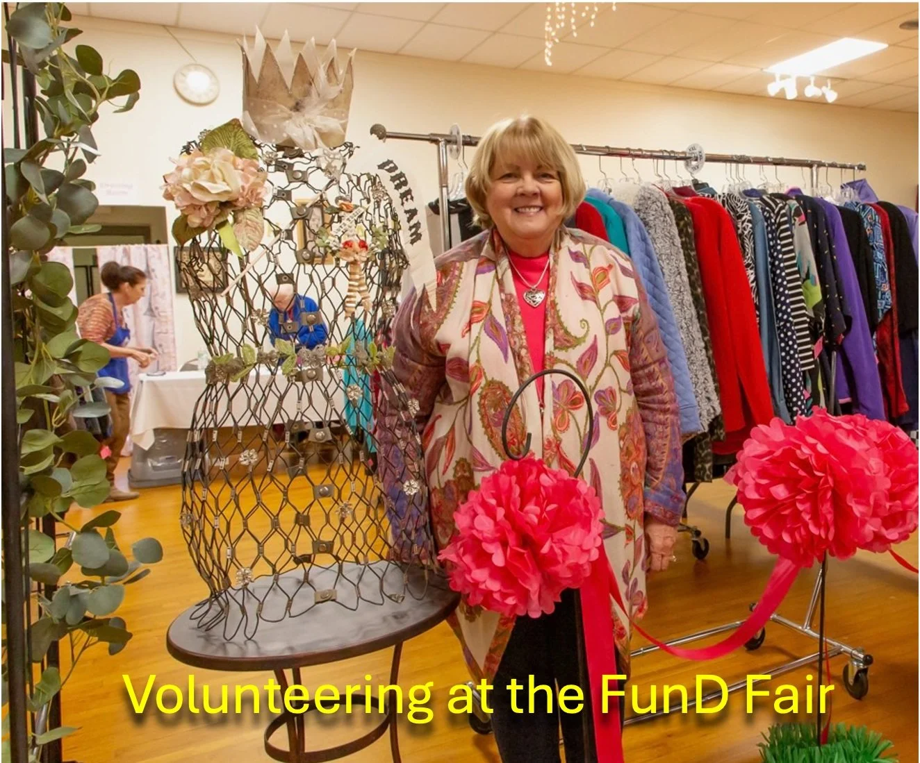 A smiling woman volunteering at a craft fair, standing next to a wire mannequin decorated with jewelry, with pink tissue paper pom-poms, a rack of colorful jackets, and other display tables in the background.