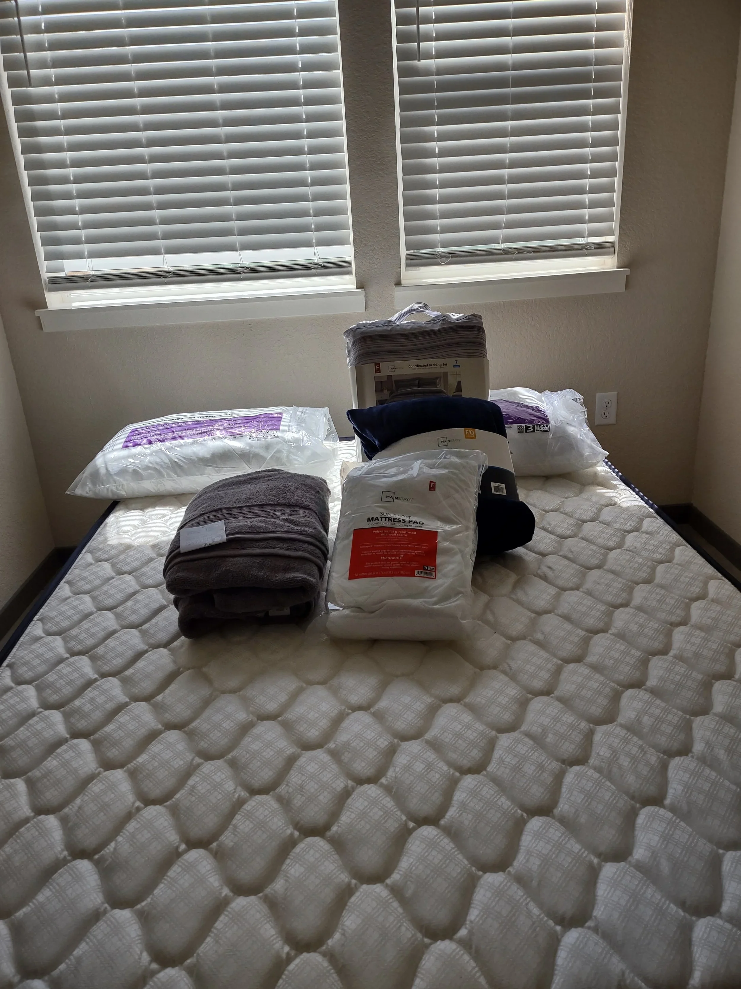 A bed with a mattress, with pillows and linens in plastic packaging, placed underneath two windows with closed blinds.