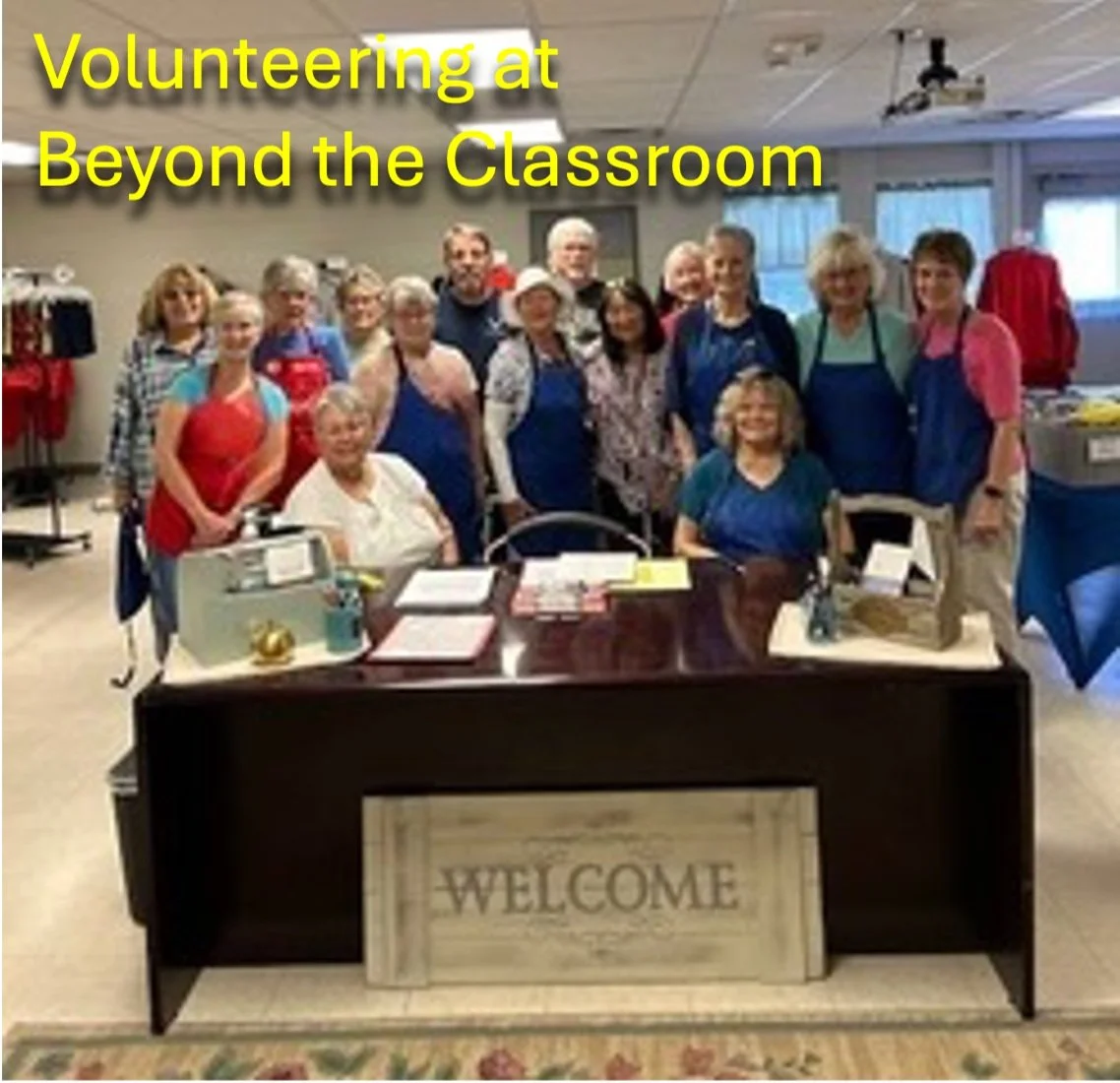 Group of volunteers standing behind a welcome desk in a classroom setting, with a mix of older adults and younger individuals, some wearing aprons.