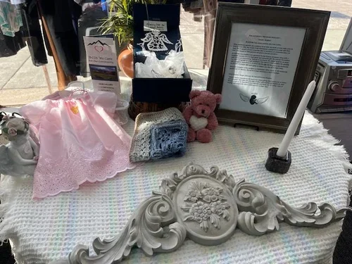 A display table with a pink cloth, a small stuffed pink bear, a framed document, a candle, and decorative white scrolls, set outdoors.