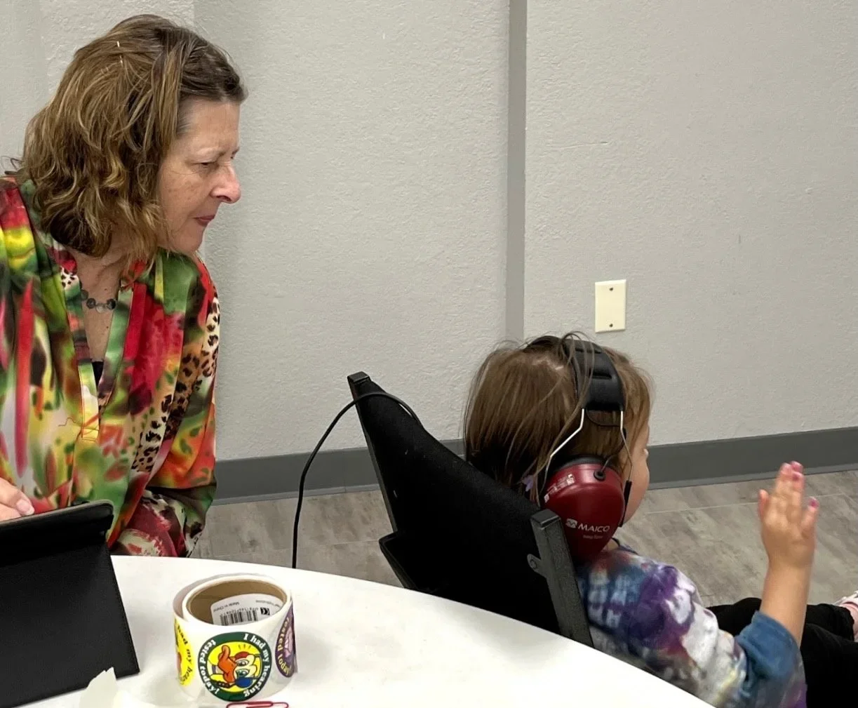 A woman with curly hair wearing a colorful, patterned shirt, conducting a hearing test with a young girl sitting in a chair, wearing large red headphones and a tie-dye shirt, in a room with plain gray walls.