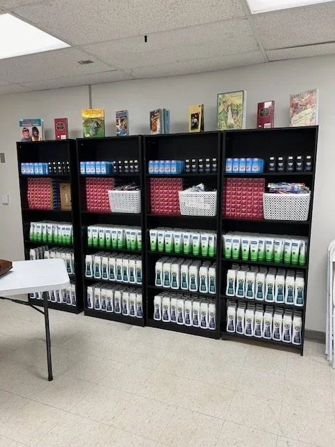 Black shelves filled with bottles of hand sanitizer, baskets, and books, with a table nearby in a classroom or office setting.