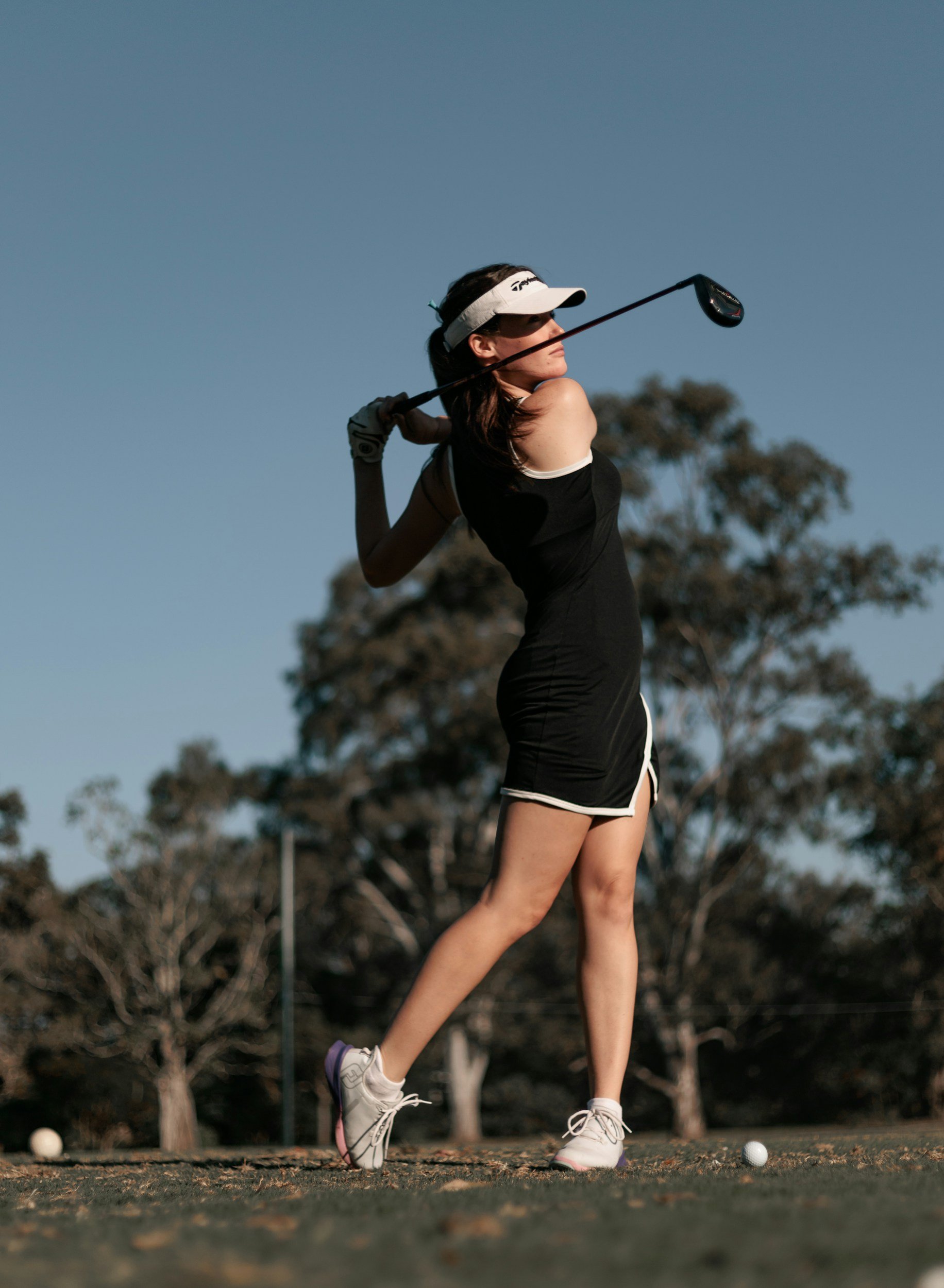 A woman playing golf on a grassy field, swinging a golf club, with trees in the background.