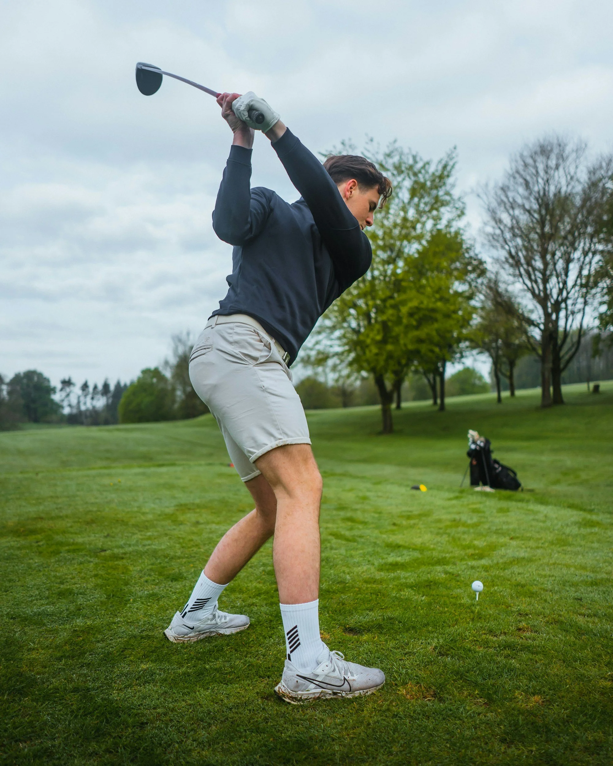Young man preparing to hit a golf ball on a golf course, wearing white shoes, white socks, beige shorts, a navy long-sleeve shirt, and holding a golf club raised behind him with trees and a bag of golf clubs in the background.