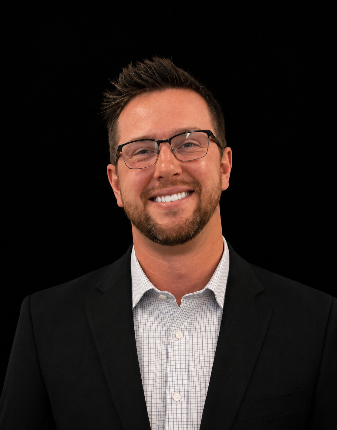 A smiling man with glasses, a beard, and short brown hair, wearing a black suit jacket and a white checkered shirt, against a black background.