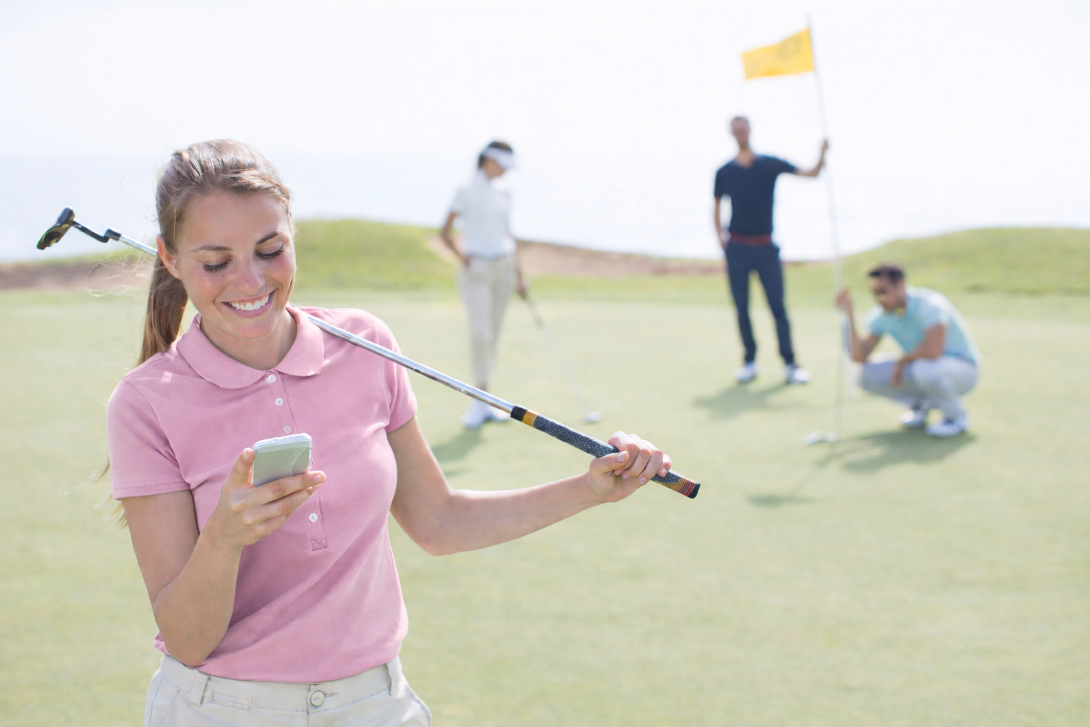 A woman smiling and looking at her phone on a golf course with three people in the background playing golf near a flagstick.