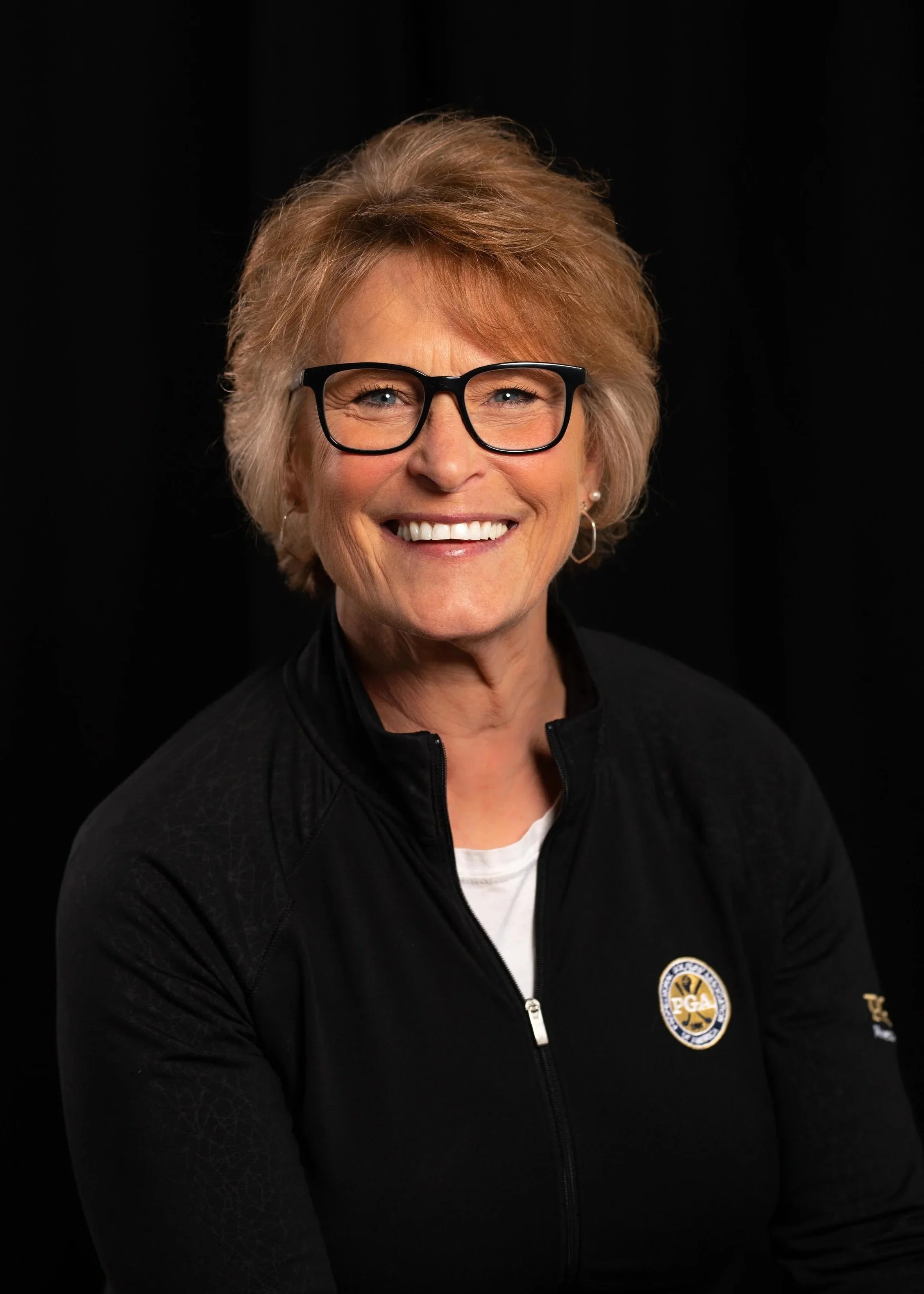 A smiling woman with short blond hair, black glasses, and pearl earrings wearing a black jacket with a PGA logo, against a black background.