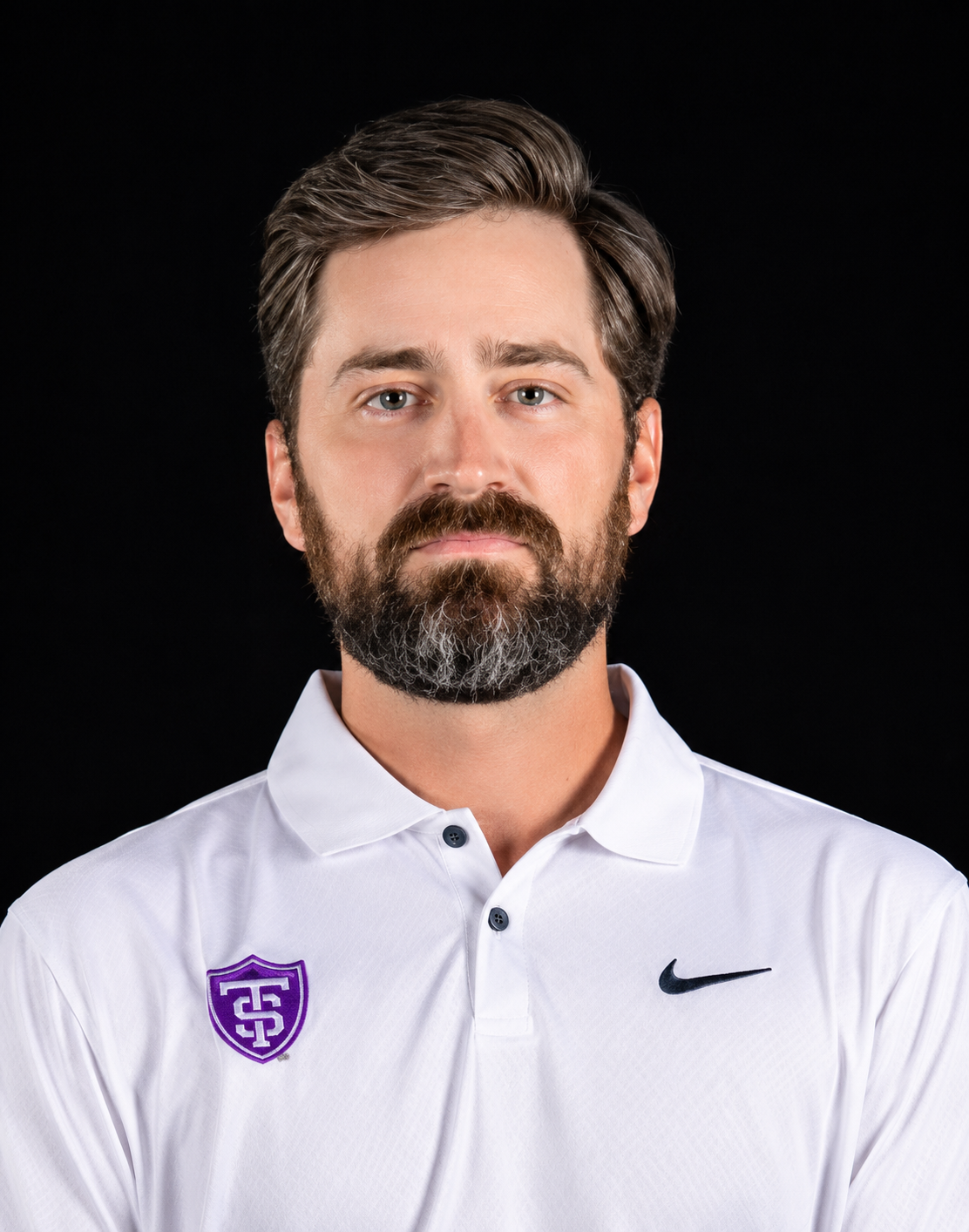 A man with a beard and mustache wearing a white polo shirt with a purple and black logo on the left side and a black Nike swoosh on the right side, against a black background.
