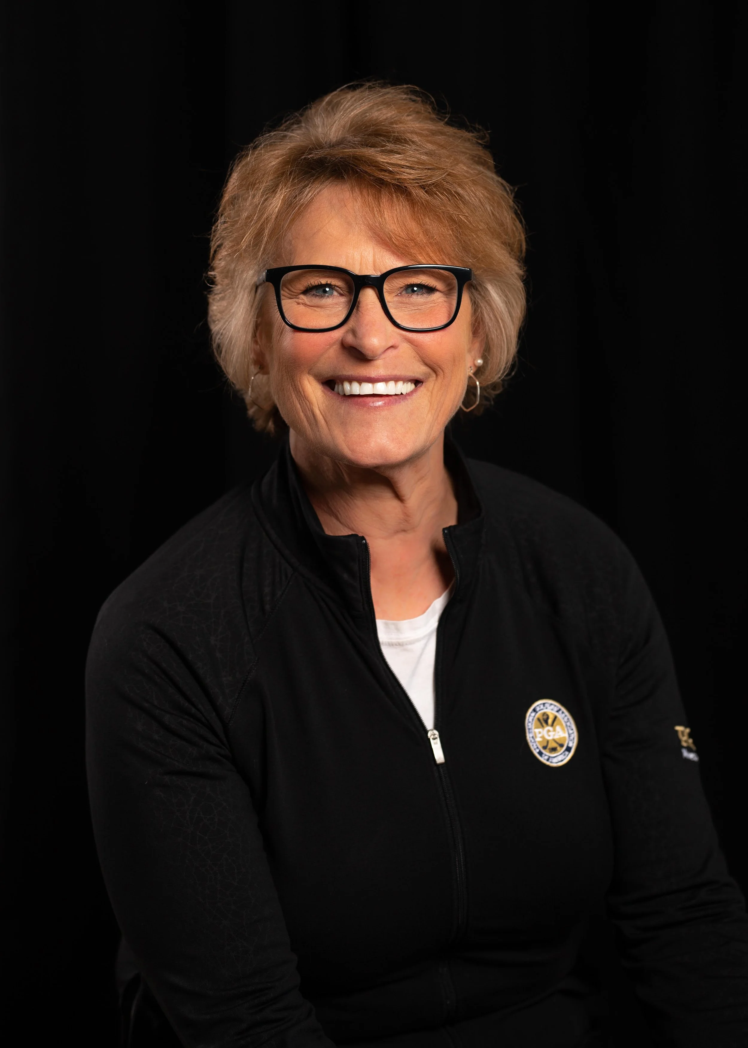 A smiling older woman with short blonde hair and black glasses, wearing a black jacket with a logo on the chest, against a black background.