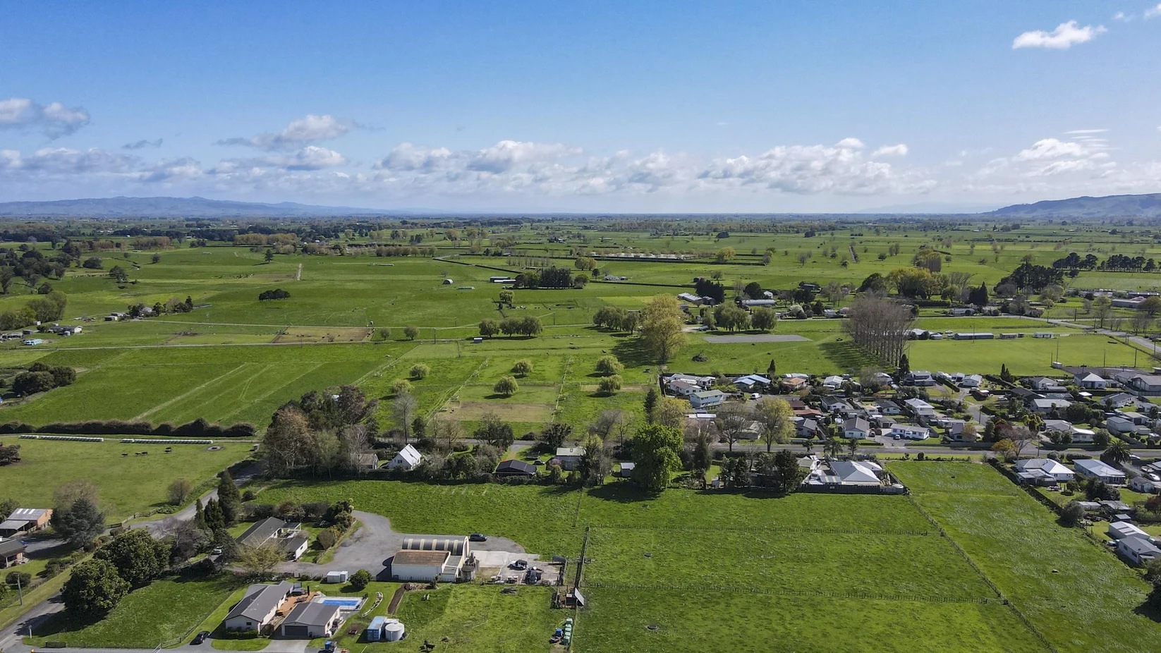 Aerial view of a rural landscape with green farmland, scattered houses, trees, roads, and hills in the distance under a partly cloudy sky.