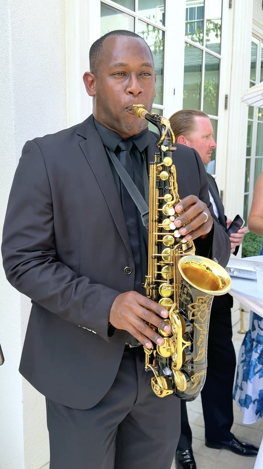 A man in a black suit playing a gold saxophone at an outdoor event.