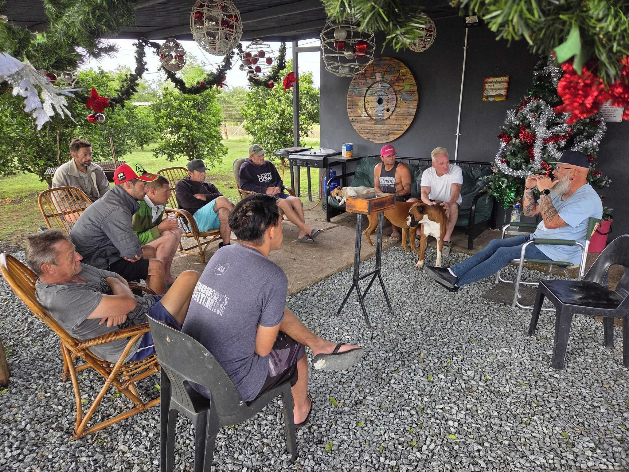 Group of people gathered outdoors, sitting and listening to a man with a beard and tattoos, wearing a hat, who is speaking or presenting. The setting is decorated with Christmas ornaments, including a Christmas tree and hanging decorations, indicating a holiday gathering.